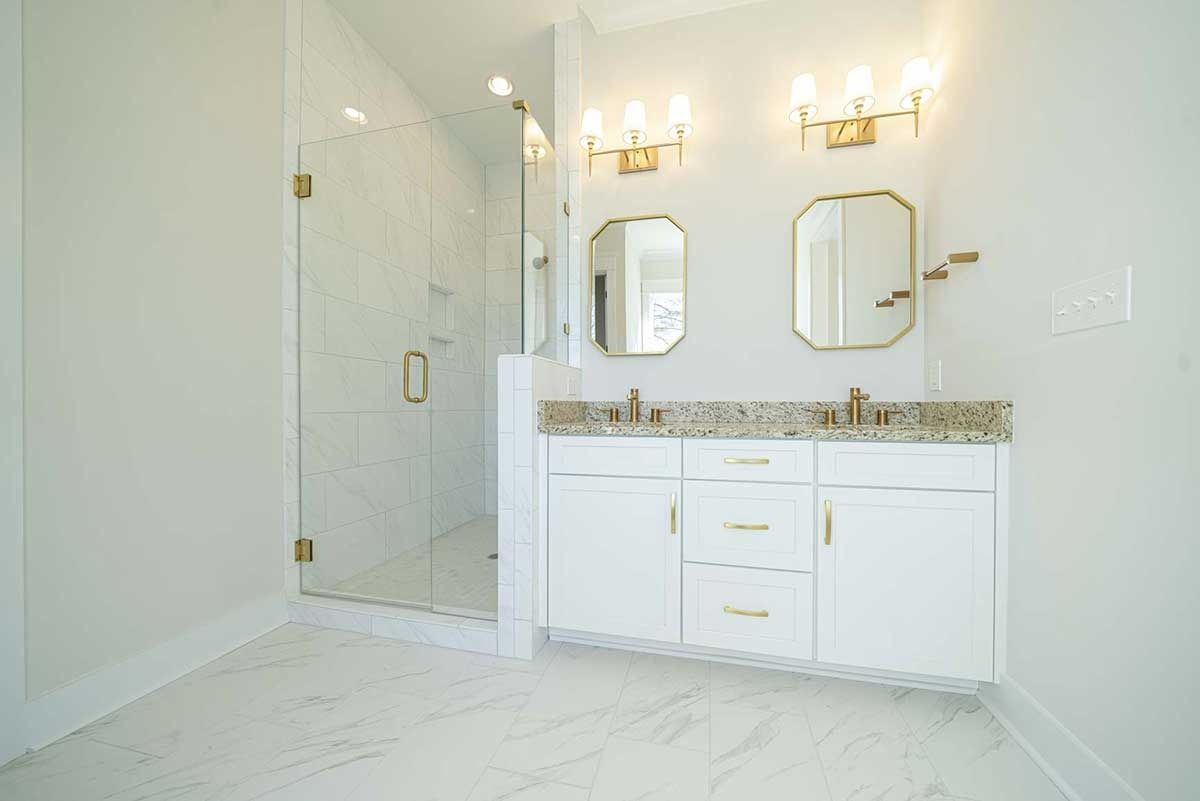 Bright white bathroom with glass shower, double vanity, gold fixtures, and marble-style tile floor.
