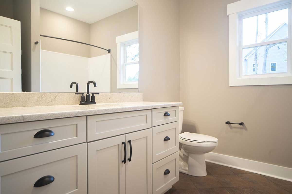 Bright bathroom with white vanity, black faucets, toilet, and two windows.
