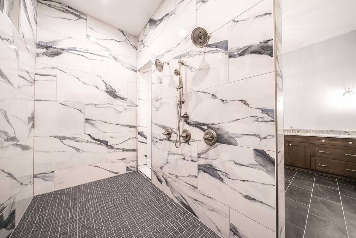 Marble-look tiled shower with dark hex floor and glass door beside a double-sink vanity