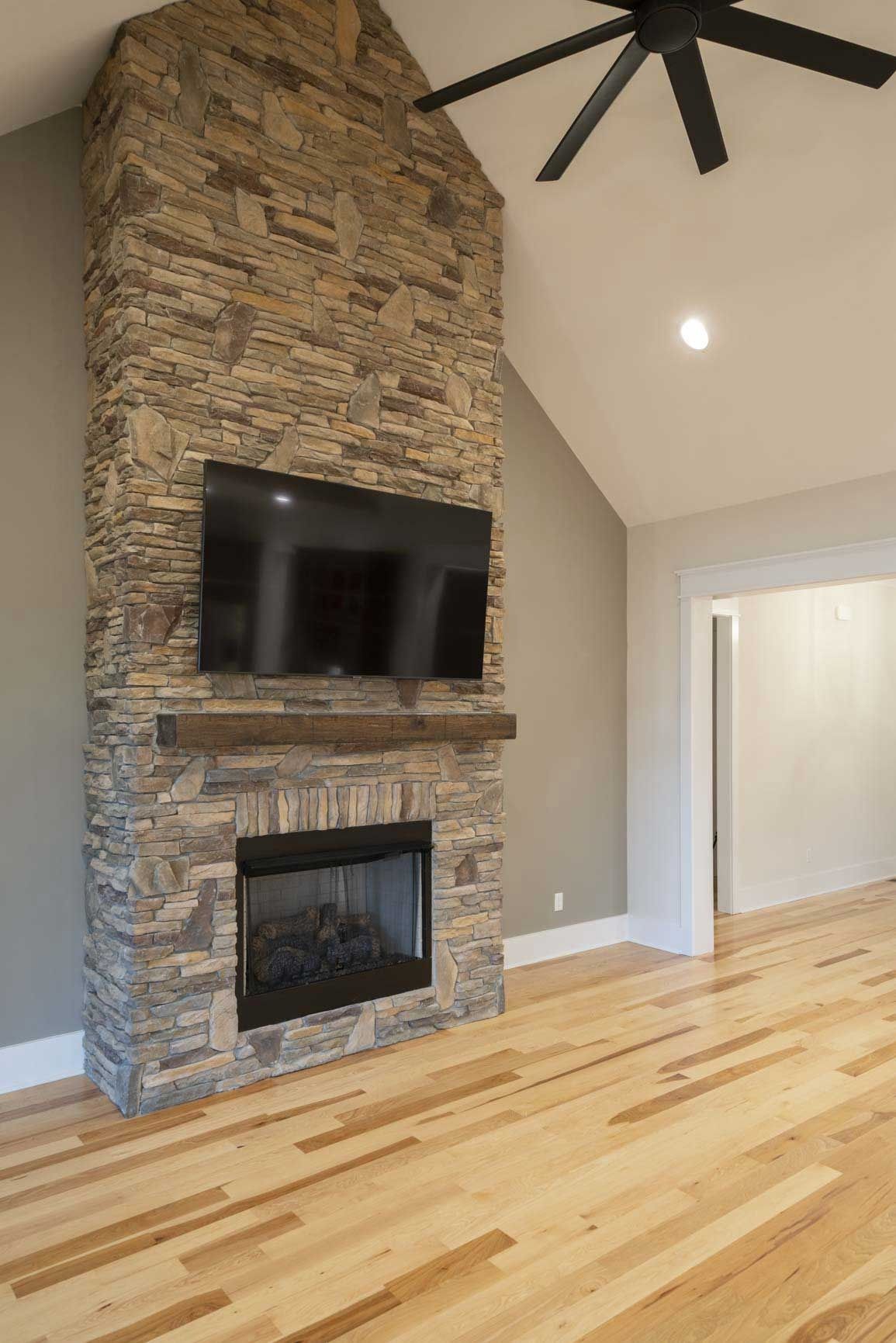 Modern living room with stone fireplace, wall-mounted TV, light wood floors, and vaulted ceiling with black fan