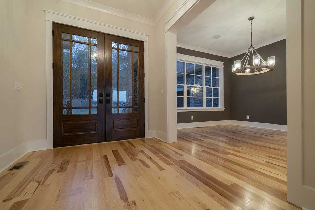 Empty living and dining room with hardwood floors, dark double front doors, and a hanging light fixture.