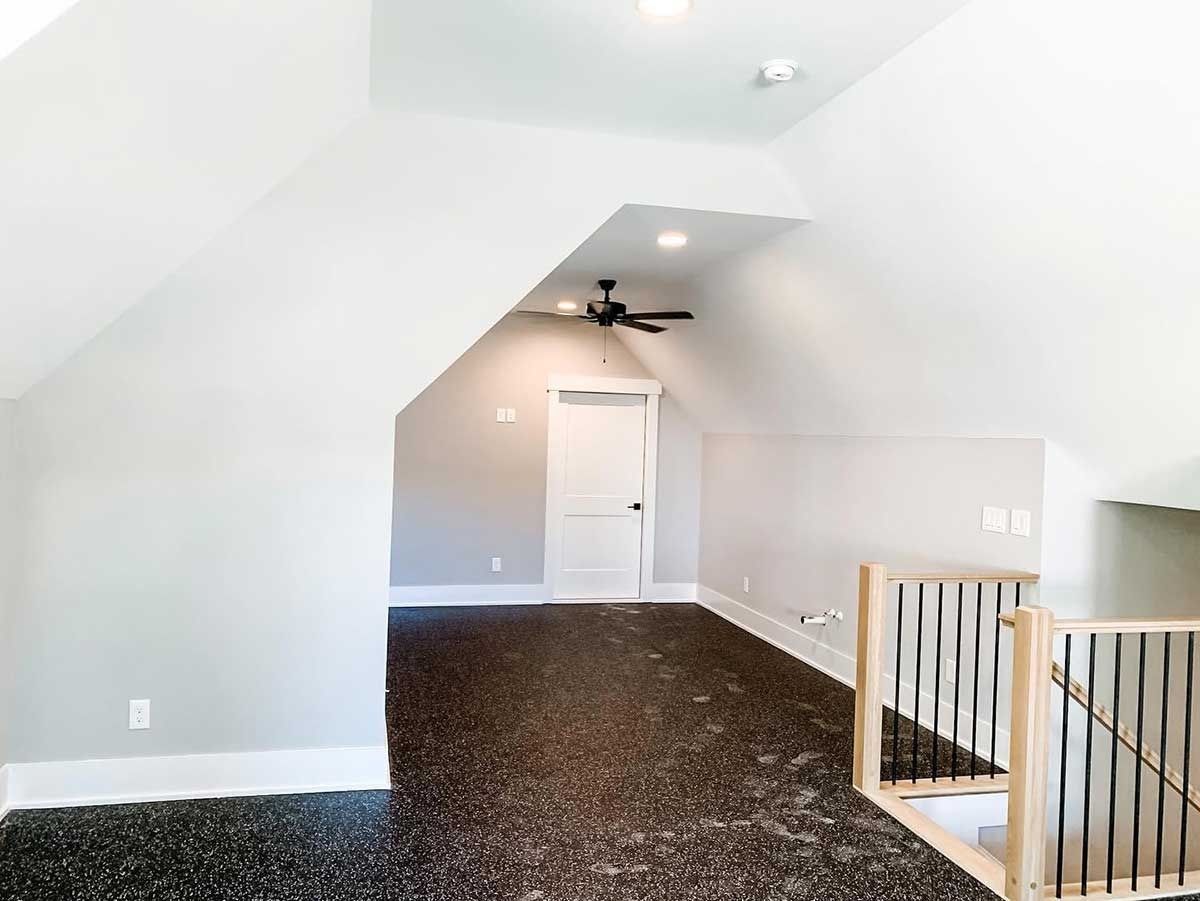 Empty attic room with sloped white walls, dark carpet, ceiling fan, and a staircase railing.