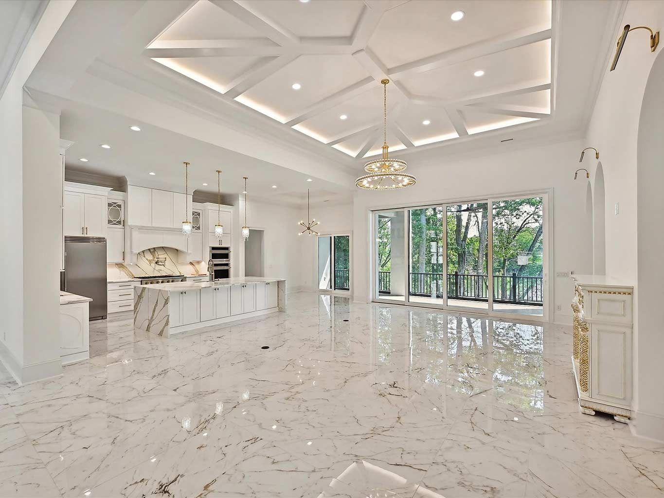 Spacious white luxury living area with marble floors, vaulted ceiling, kitchen island, and large glass doors to greenery