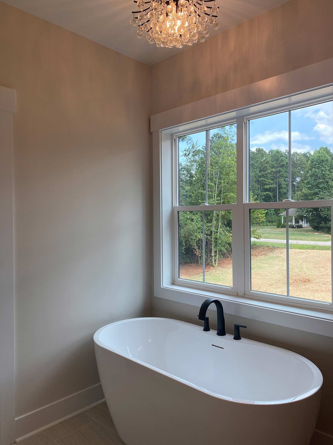 Small bathroom with a white freestanding tub beneath a large window overlooking trees and a yard.