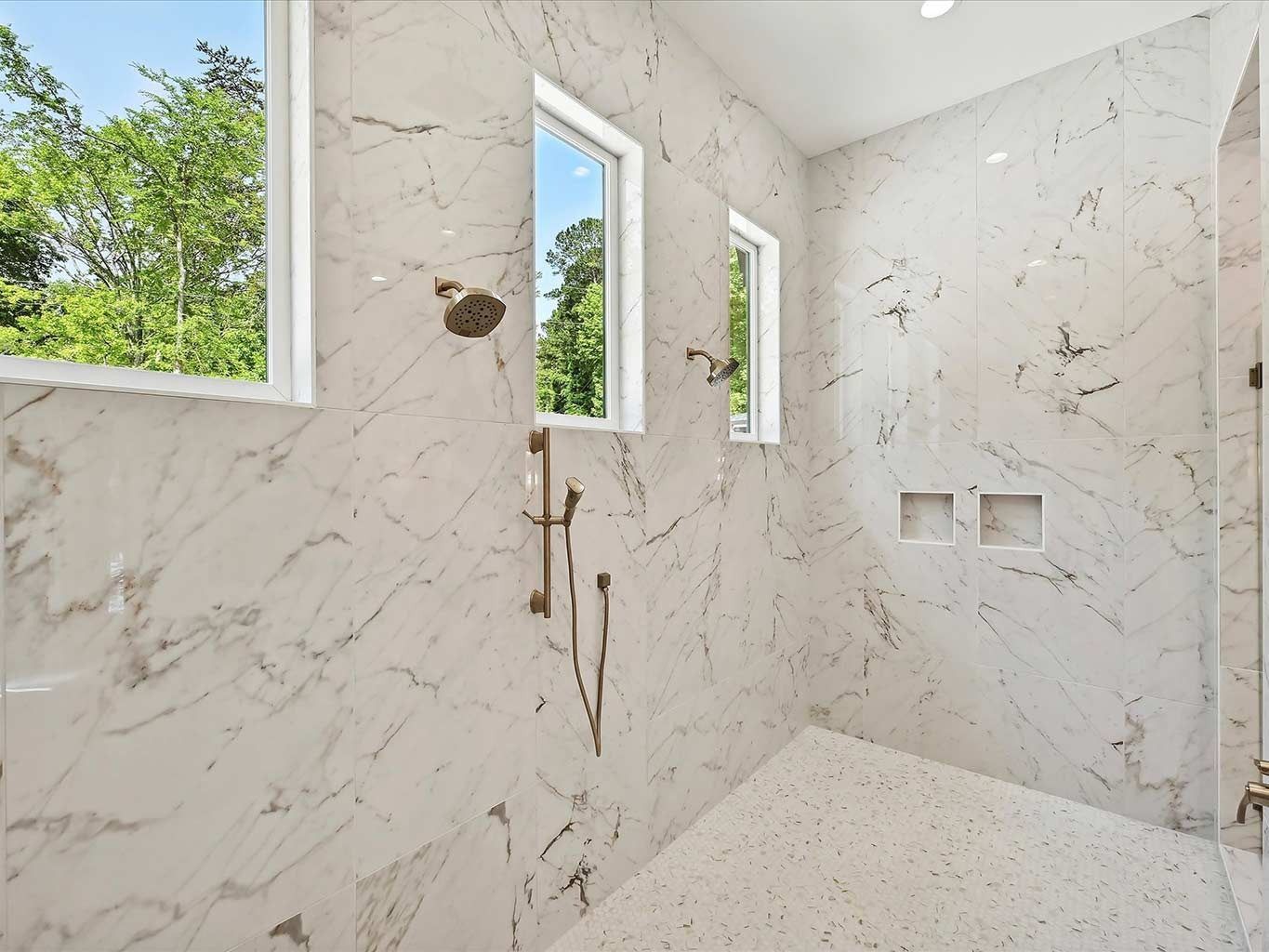 Empty marble-tiled shower with three narrow windows and chrome fixtures, overlooking greenery outside