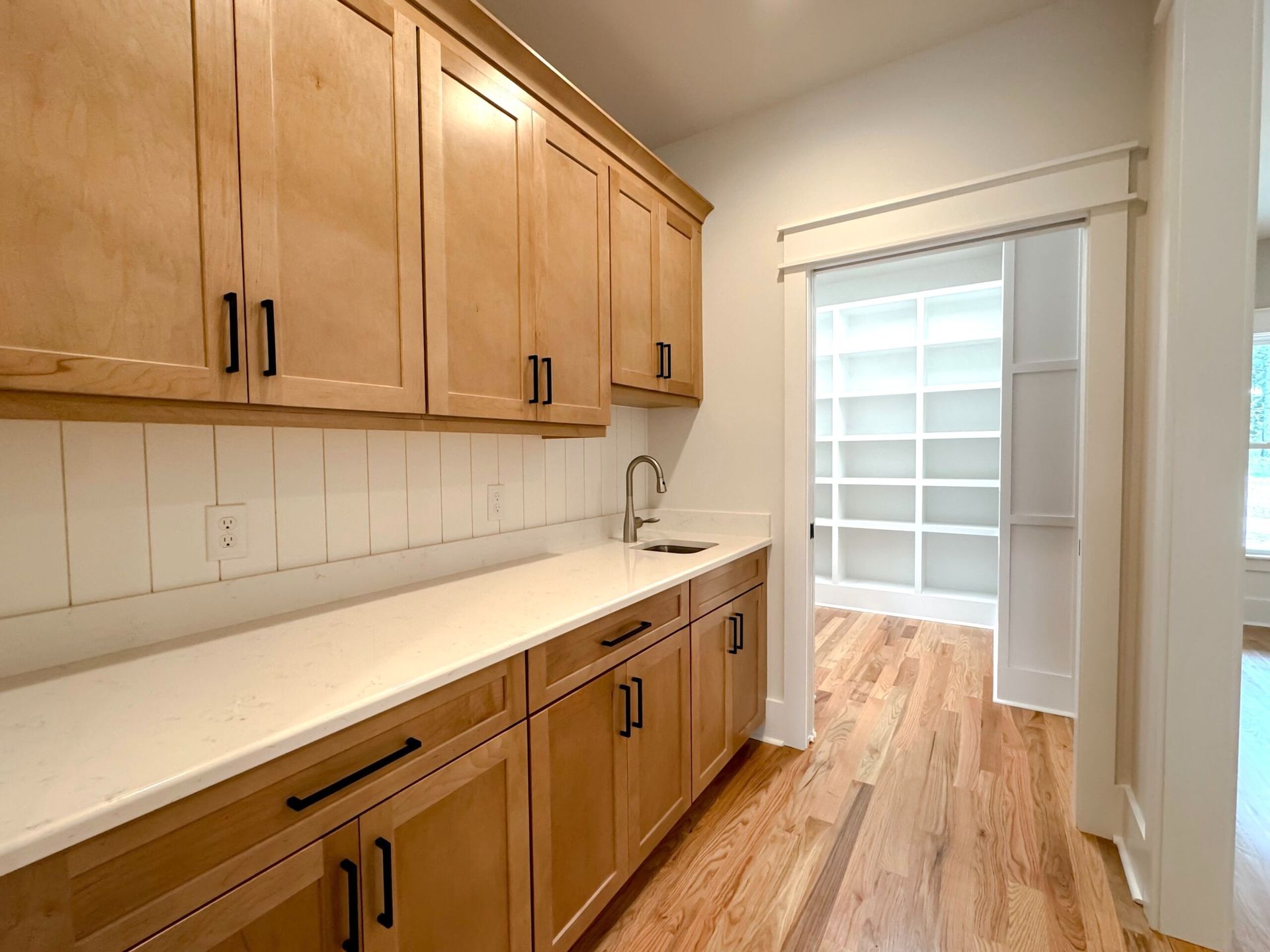 Empty kitchen with light wood cabinets, white counters, and a bright walkway to a tiled room.