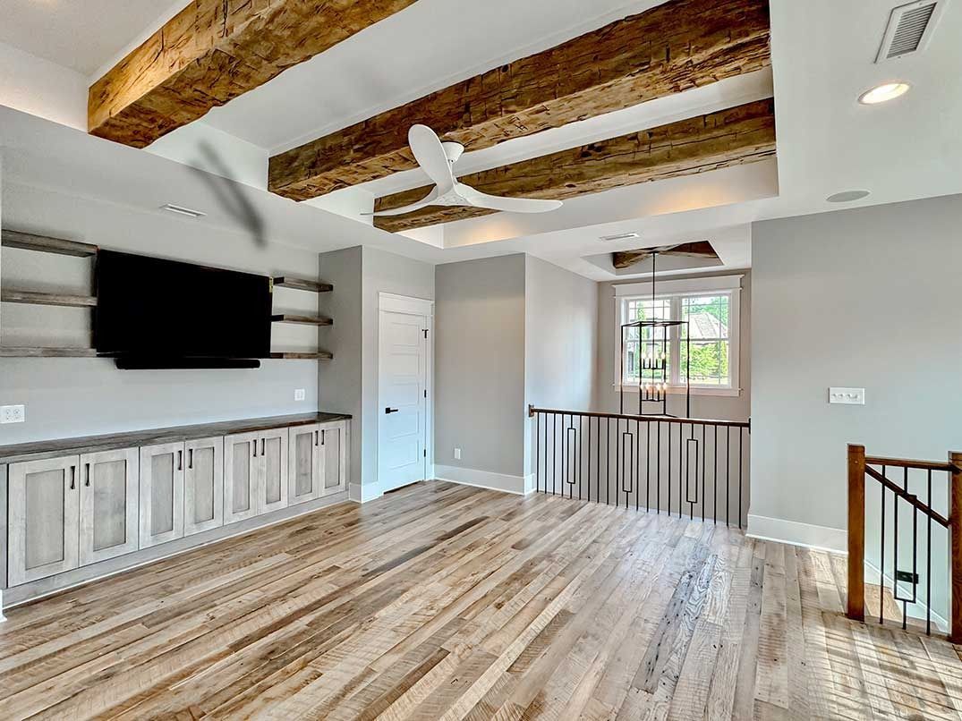 Modern empty living room with exposed wood ceiling beams, wall-mounted TV, and large window with railing.