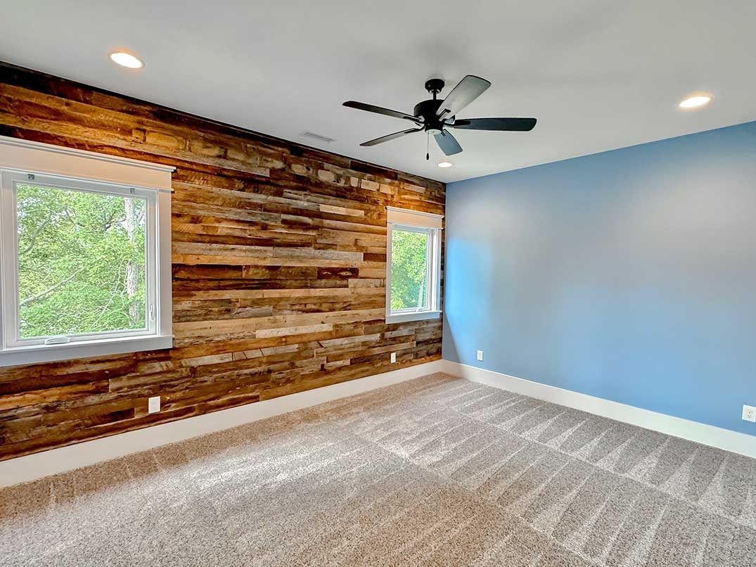 Empty room with blue walls, rustic wood accent wall, carpet, windows, and ceiling fan.