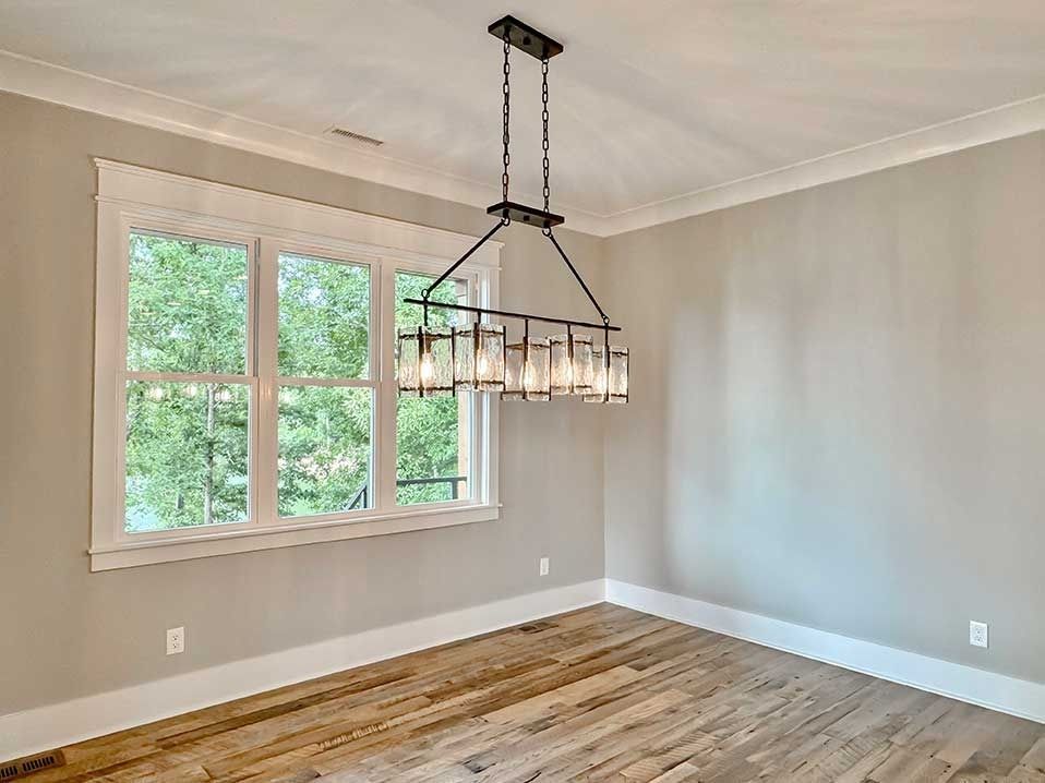Empty dining room with hardwood floors, beige walls, and a black pendant chandelier by a window.