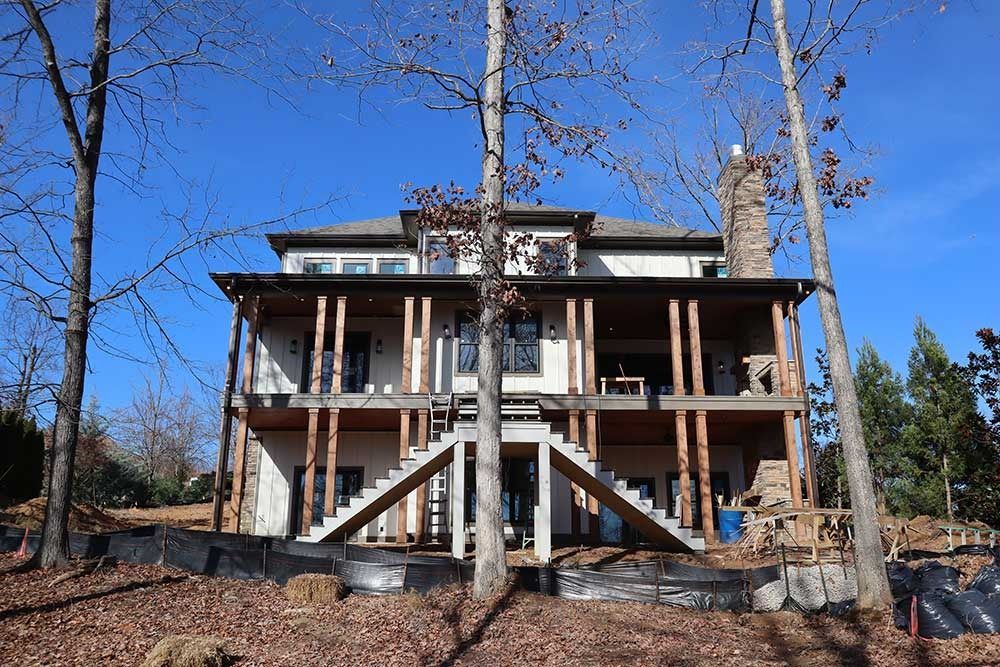 Two-story house under construction with exposed framing and a front porch, surrounded by trees
