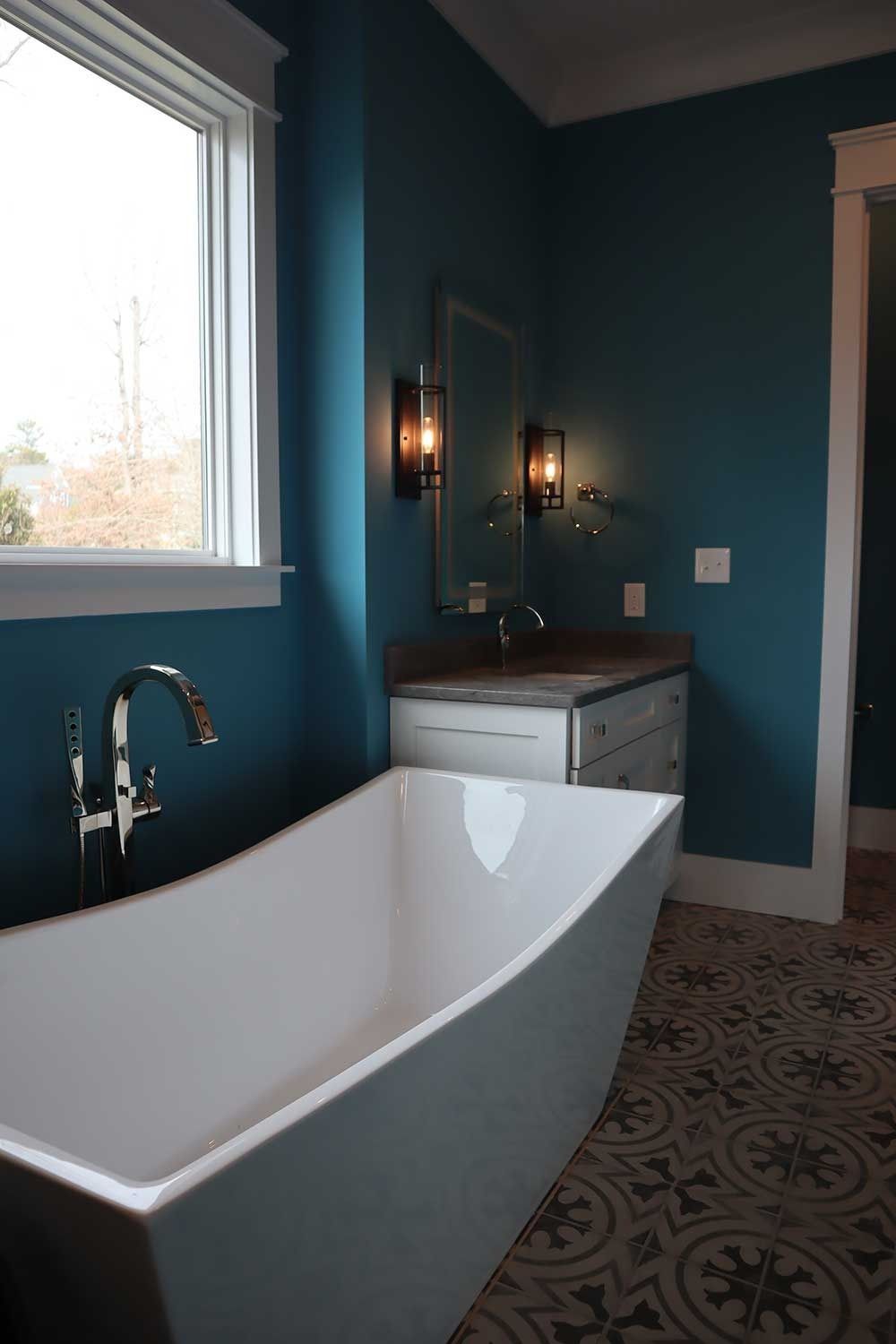 Modern bathroom with a freestanding tub, teal walls, patterned tile floor, and a window above the tub