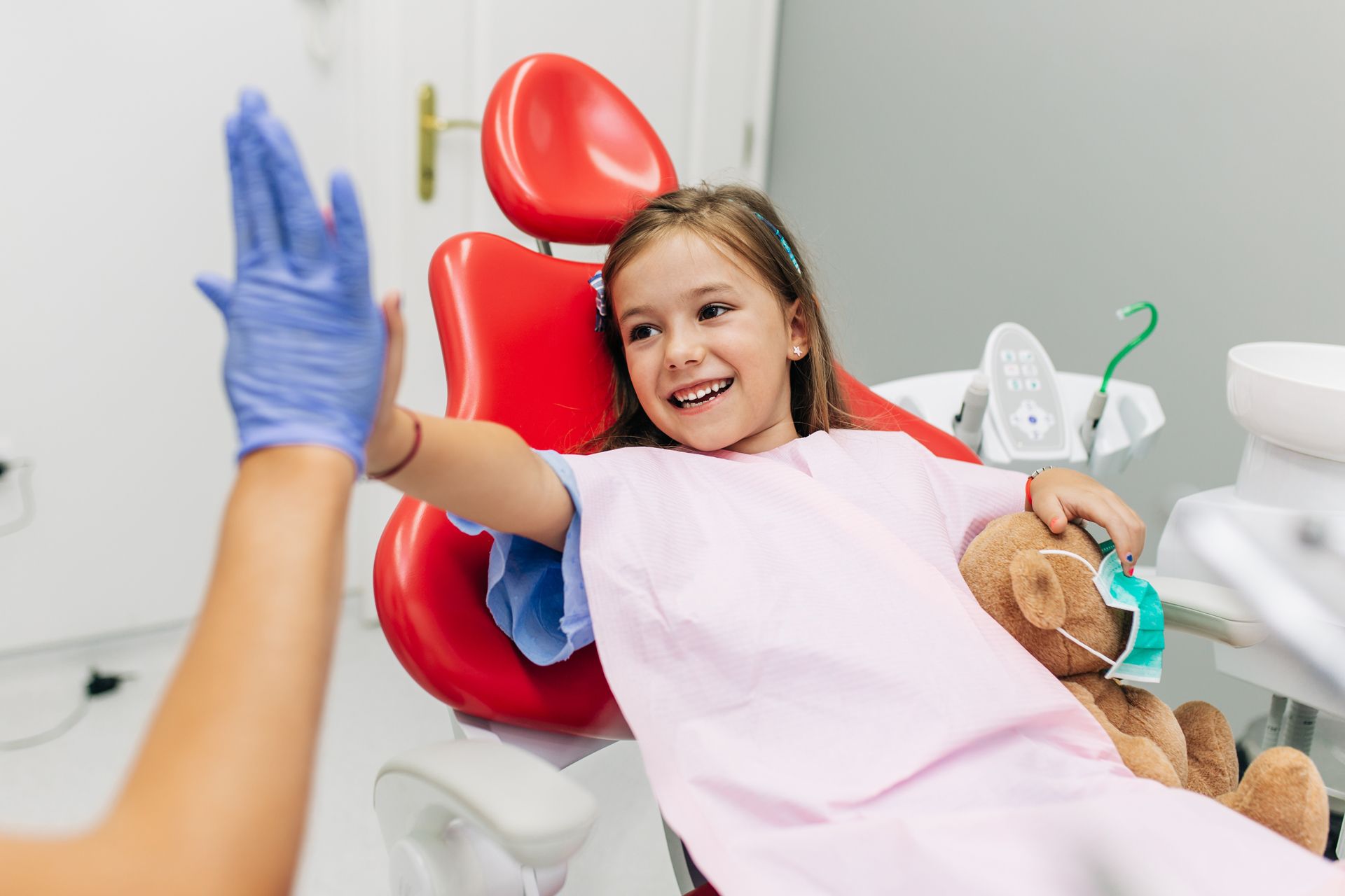 Girl in a dentist chair smiles, gives a high five. Teddy bear present. 