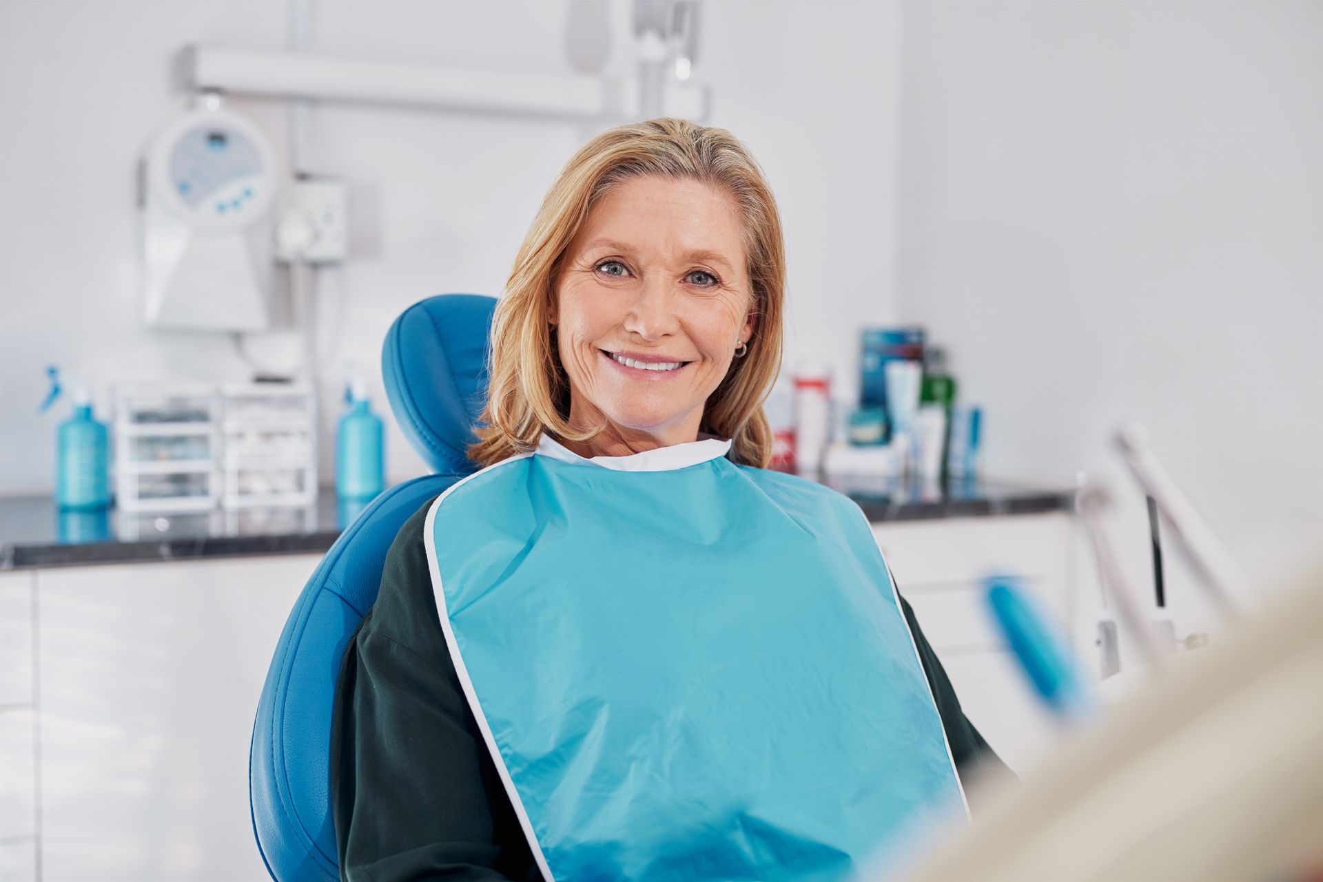 Woman smiles in a dentist's chair, wearing a blue bib in a dental office.