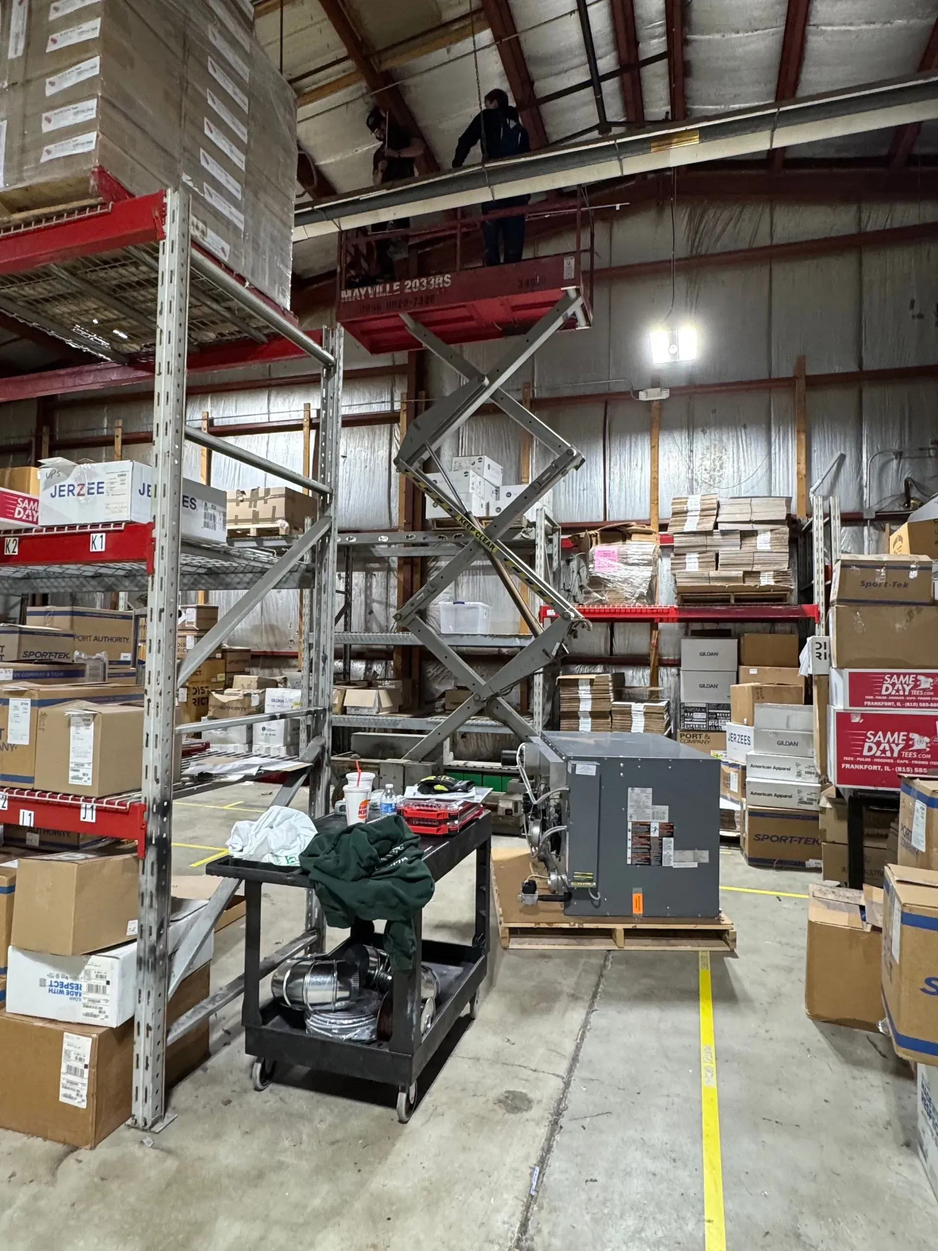 Person on scissor lift working on ceiling in warehouse, surrounded by shelves and boxes.
