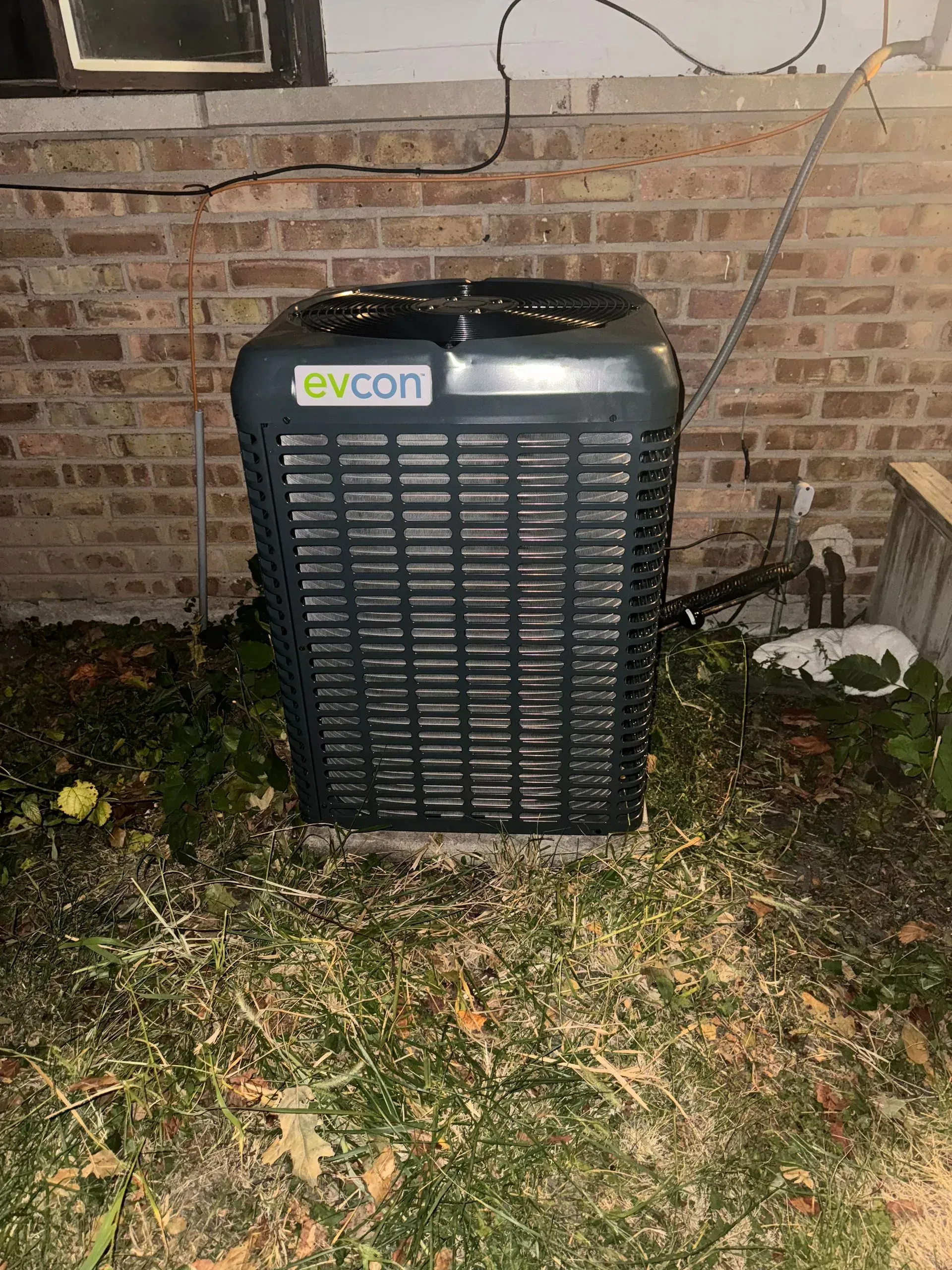 An air conditioning unit against a brick wall, surrounded by grass and debris.