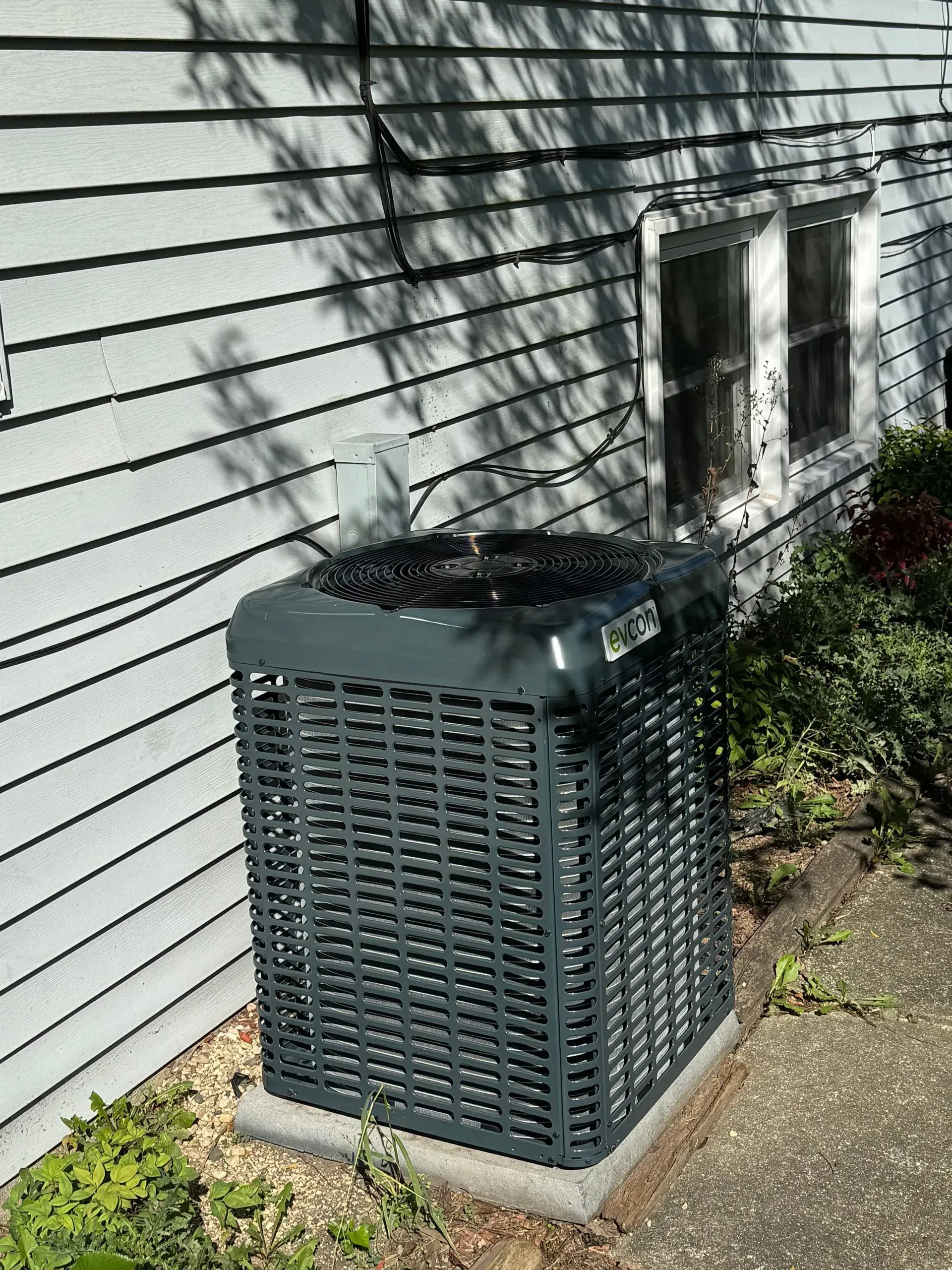 Gray air conditioning unit outside a building with gray siding, next to a window and bushes.