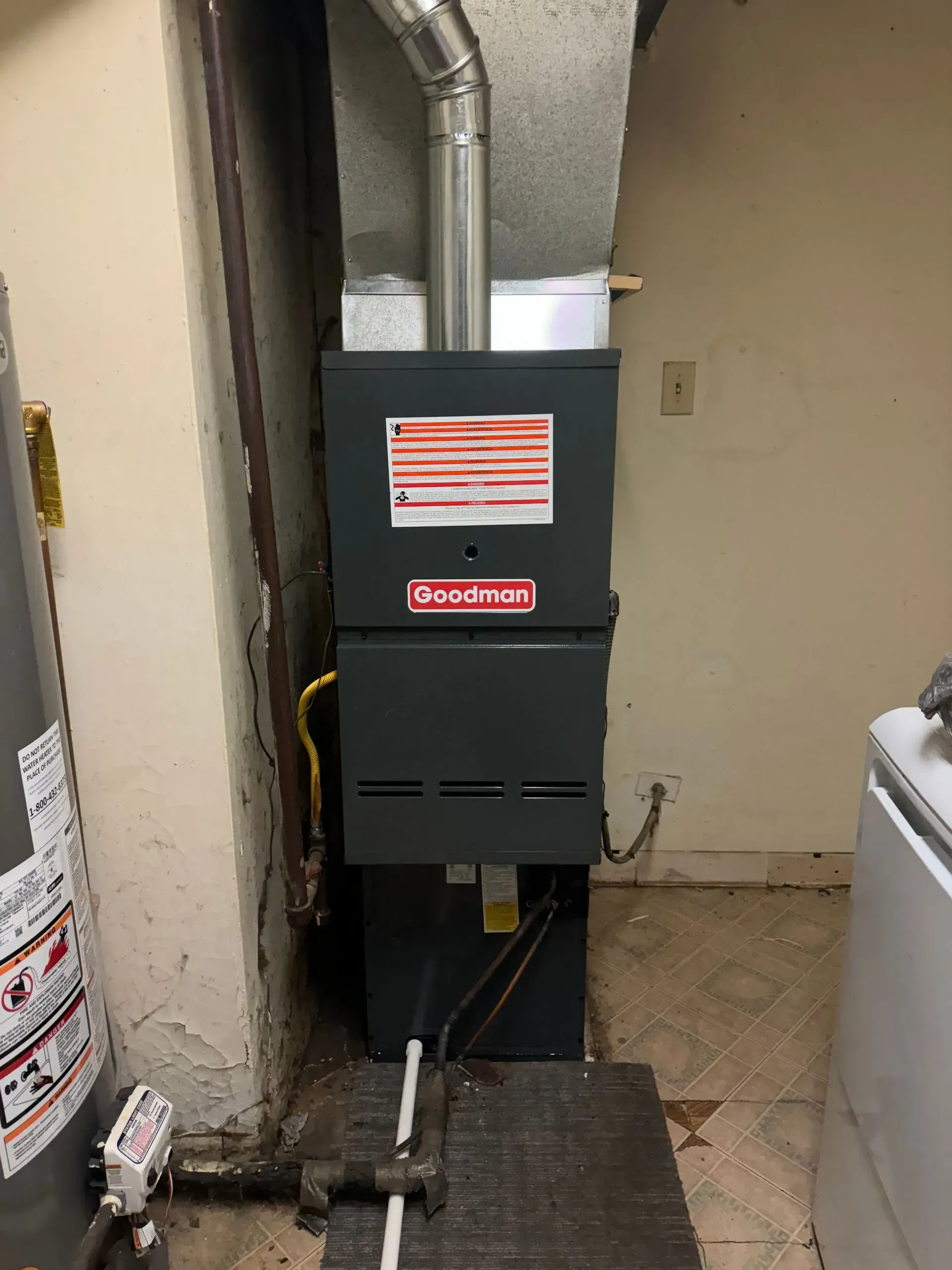 A dark gray Goodman furnace with a silver flue is in a basement. Beside it is a water heater and a washing machine.