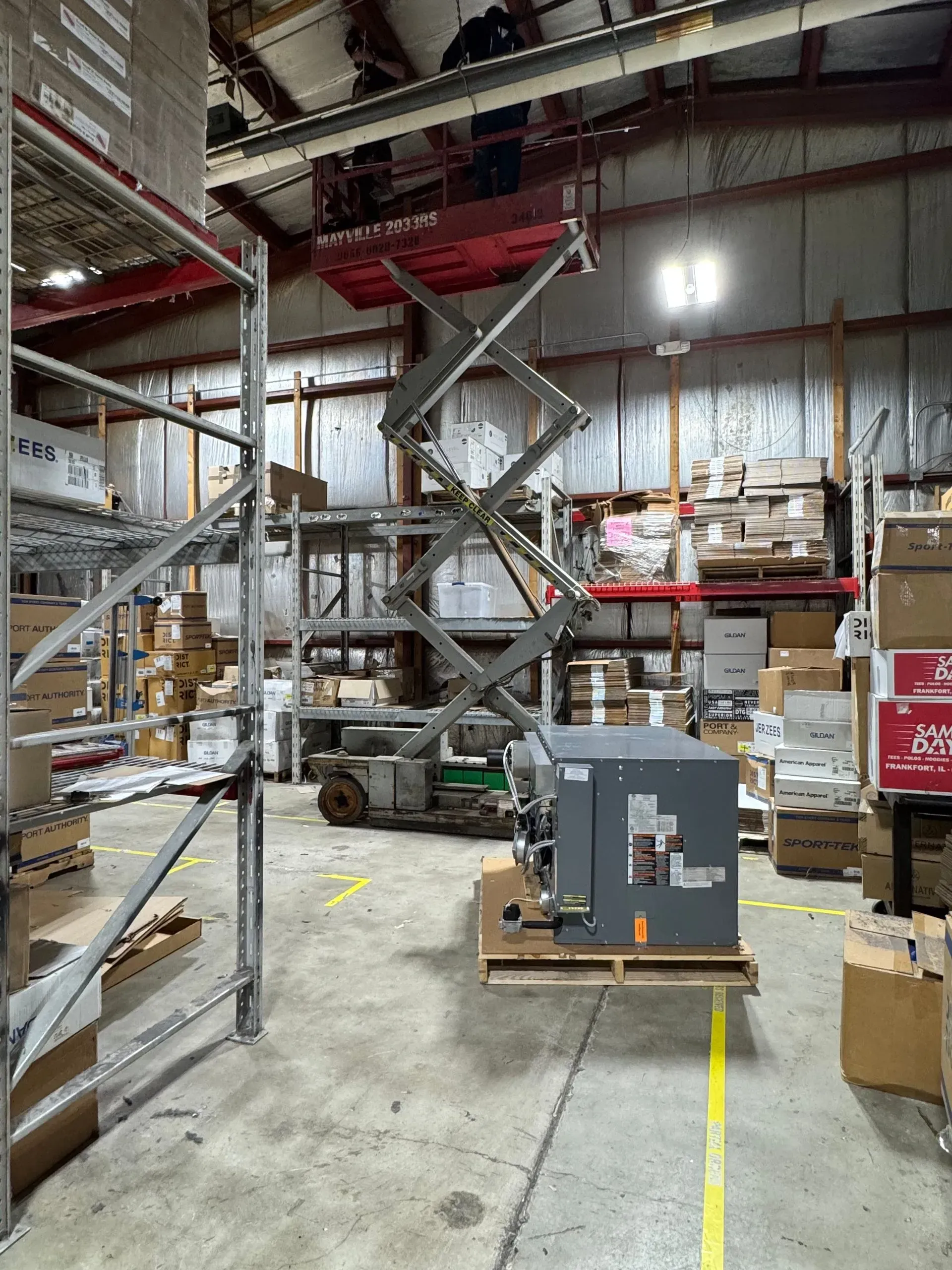 Warehouse interior with a scissor lift, shelving, and a large air conditioning unit on a pallet.