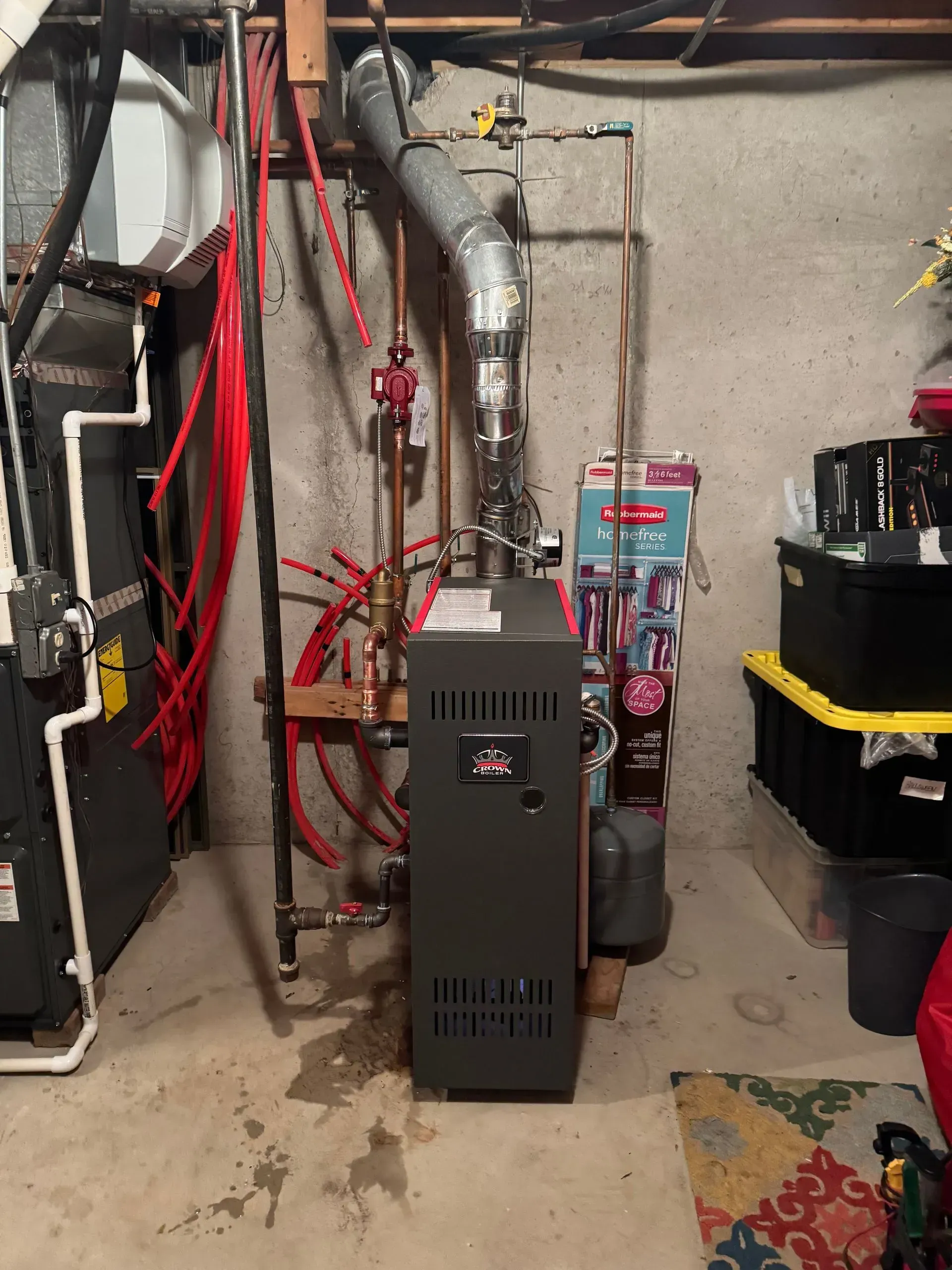 Furnace in a basement; gray appliance with various pipes, vents, and red tubing against a concrete wall.