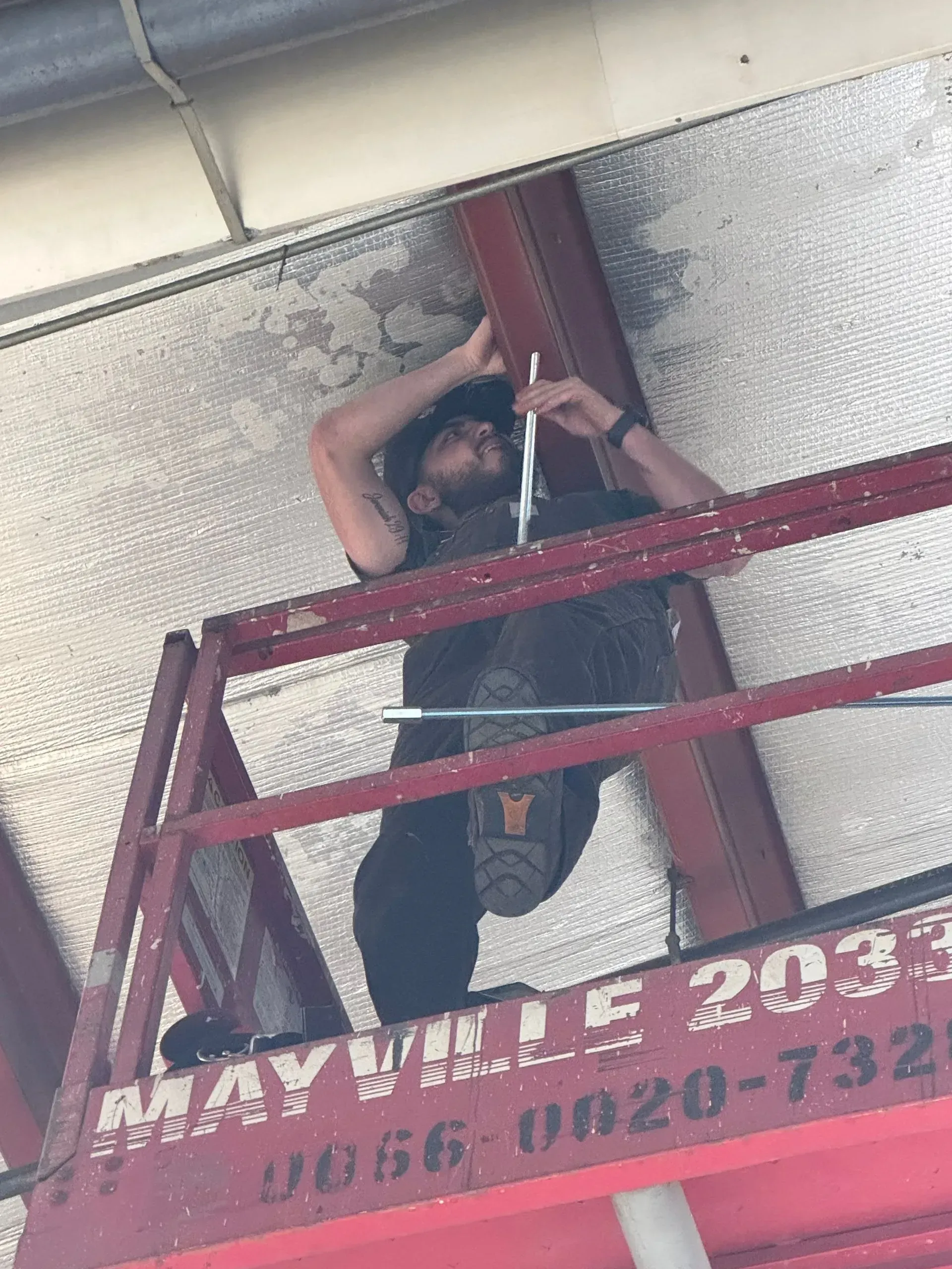 Man on a lift installing something on an overhead beam. Metallic insulation and red lift are visible.
