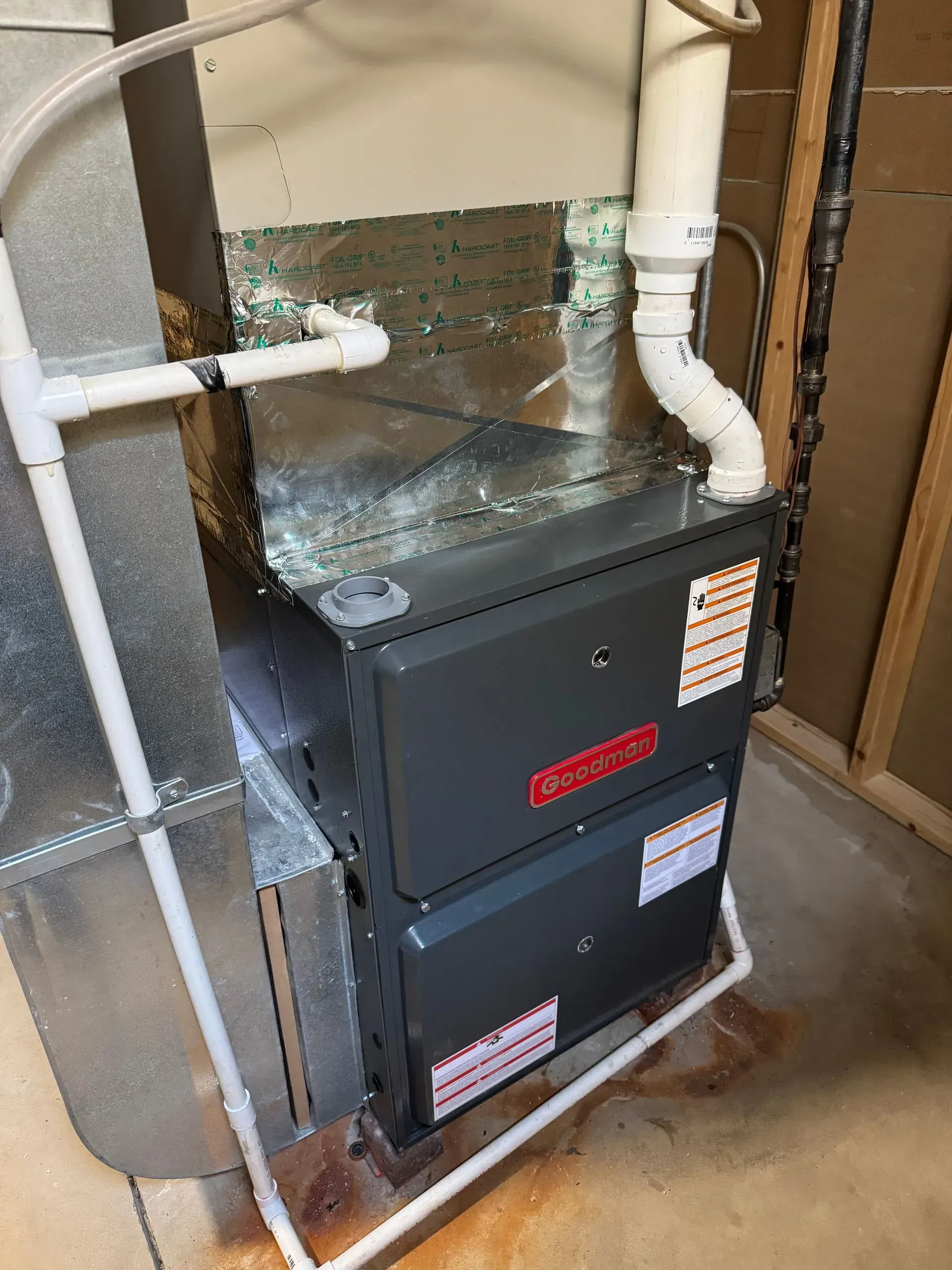 Gray Goodman furnace with white pipes, metal ductwork, and water stains in a utility room.