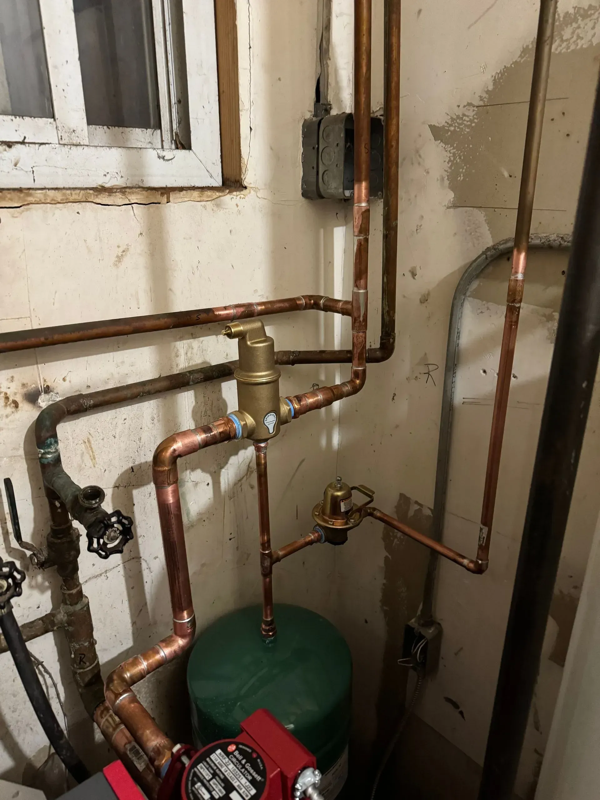 Copper pipes and fittings connected to a green tank, red pump, and a brass valve, in a basement setting.