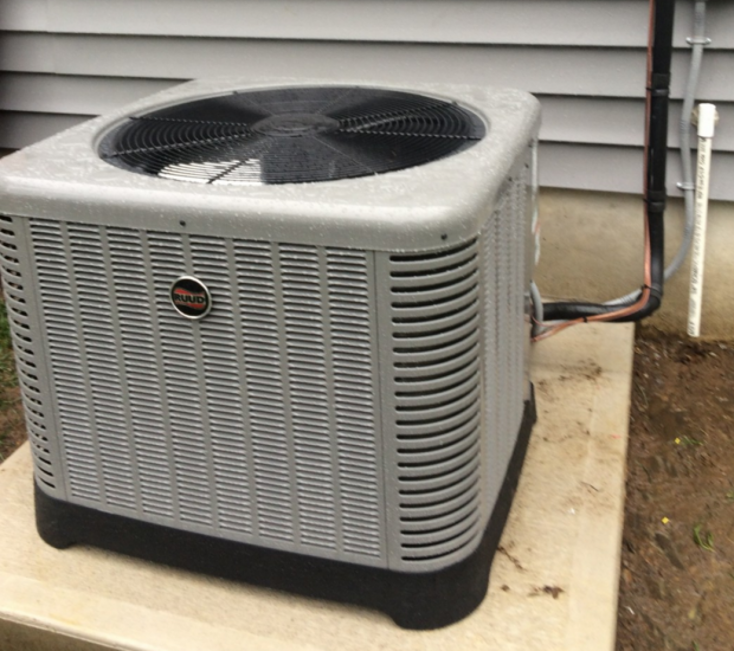A gray and black air conditioner is sitting on a concrete platform outside of a house.