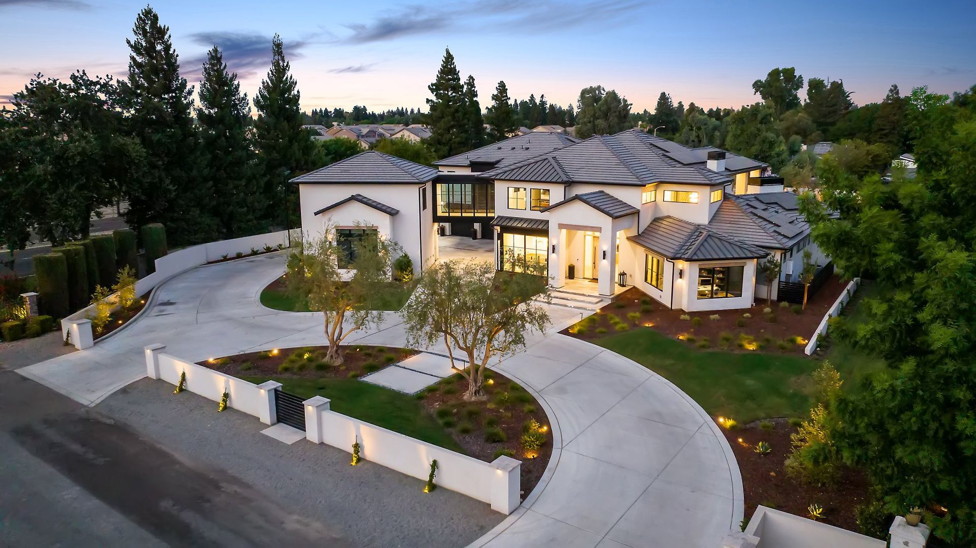Aerial view of a large modern house with a curved driveway at dusk, surrounded by trees and landscaped grounds