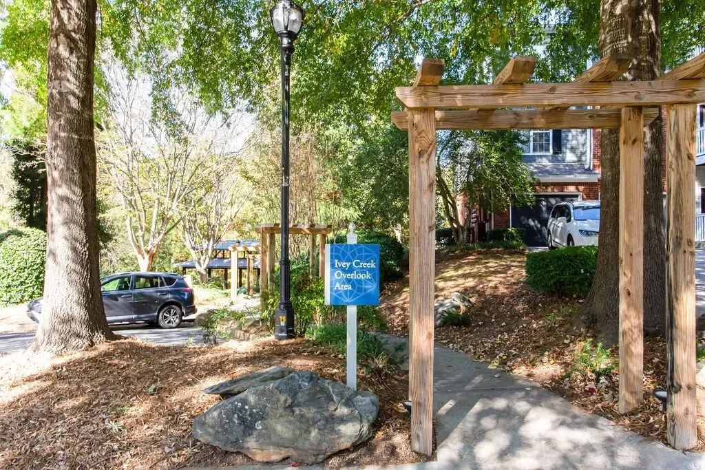 Wooded park path with a blue sign, trees, rocks, and a wooden pergola entrance.