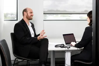 Two people in business attire meeting across a desk with a laptop in a bright office
