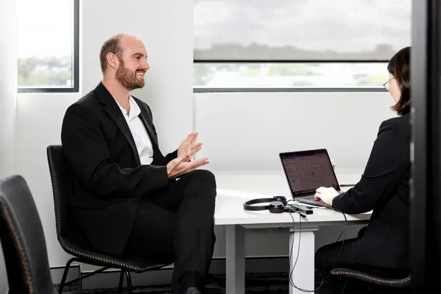 Two people in business attire meeting across a desk with a laptop in a bright office