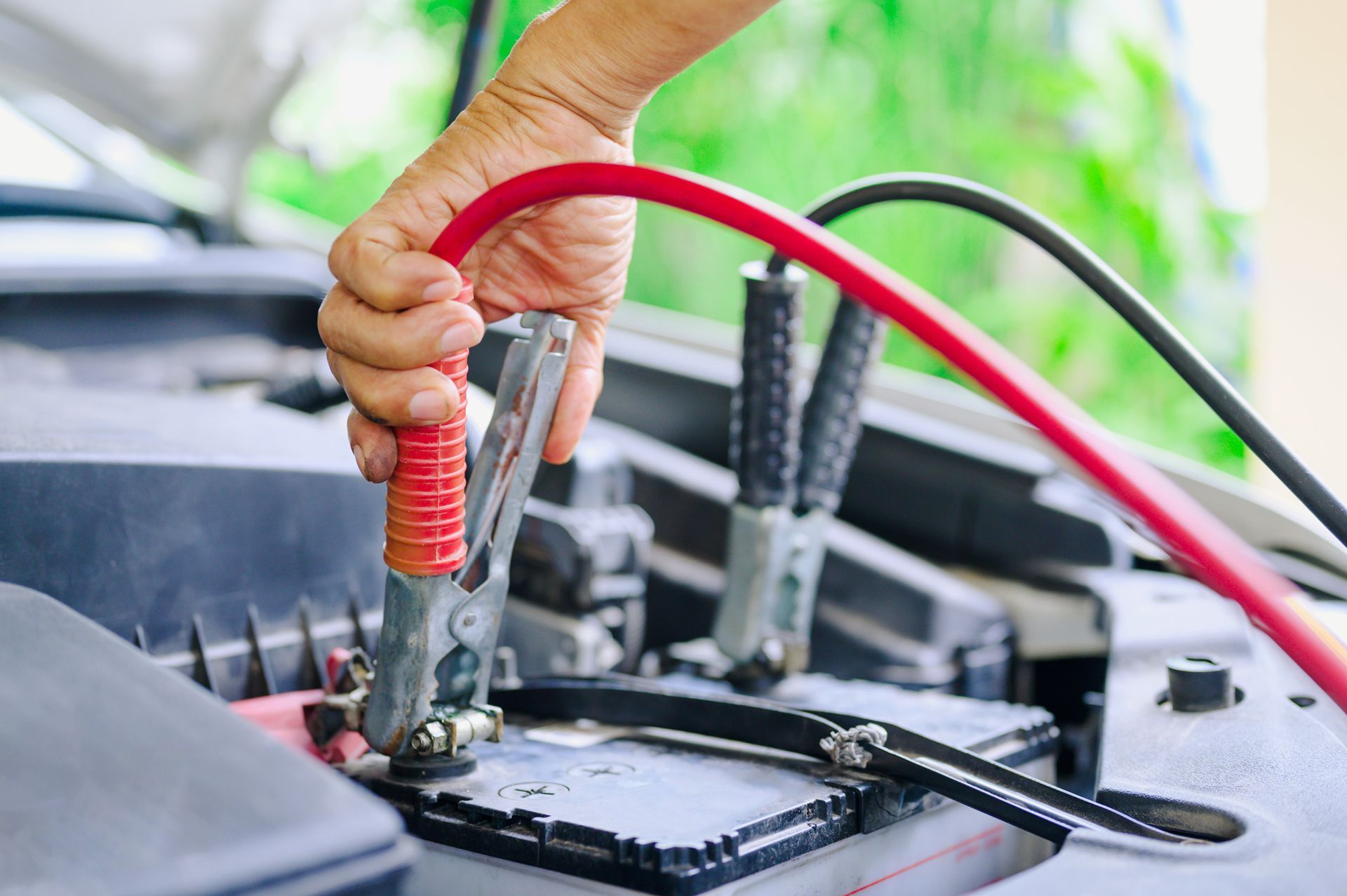 Hand connecting a red jumper cable to a car battery.