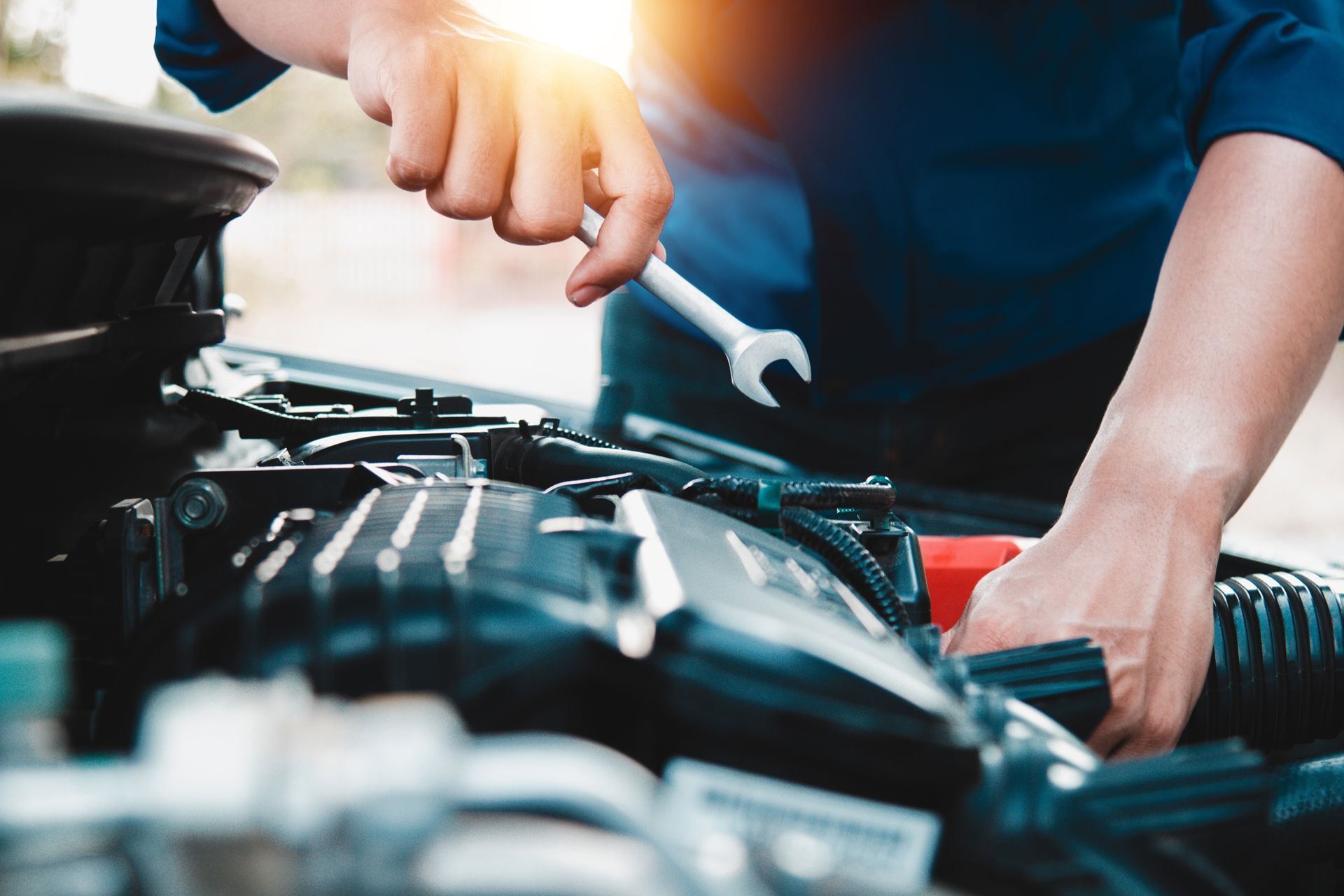 Person uses a wrench to work on a car engine, outdoors. Bright sunlight.