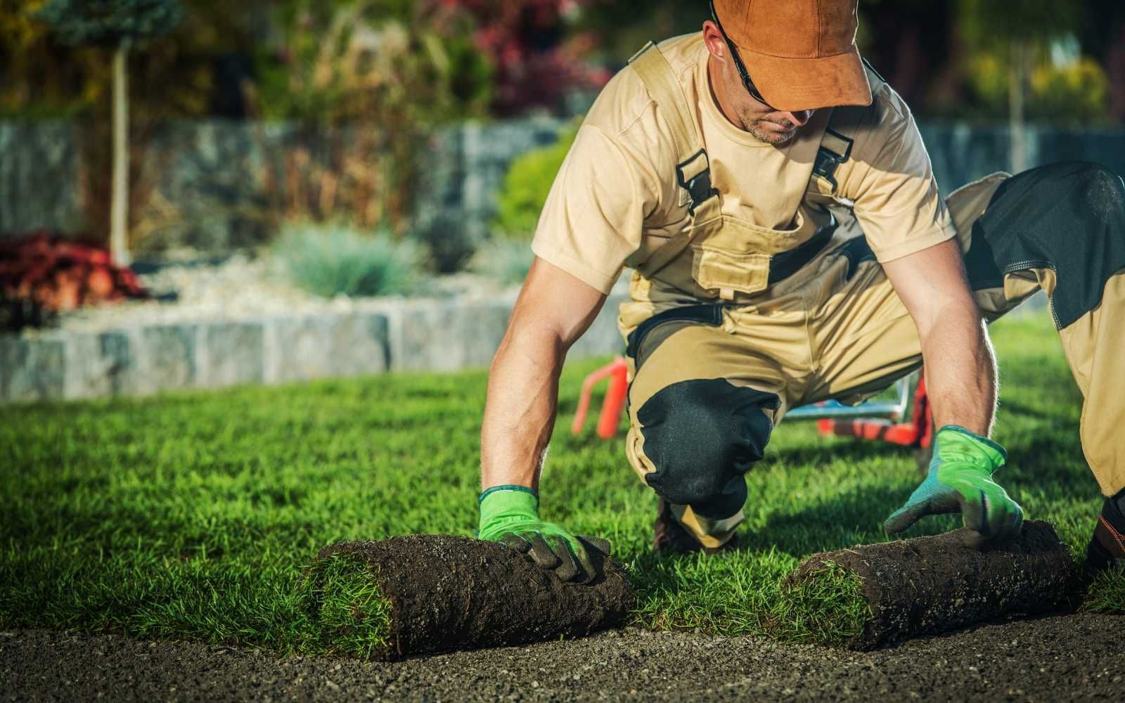 A person in work clothes is carefully laying down a roll of sod onto a lawn.