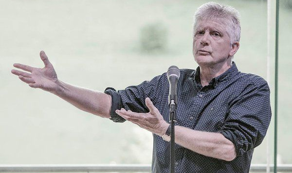 A person wearing a dark patterned shirt stands at a microphone, gesturing with both hands while speaking.