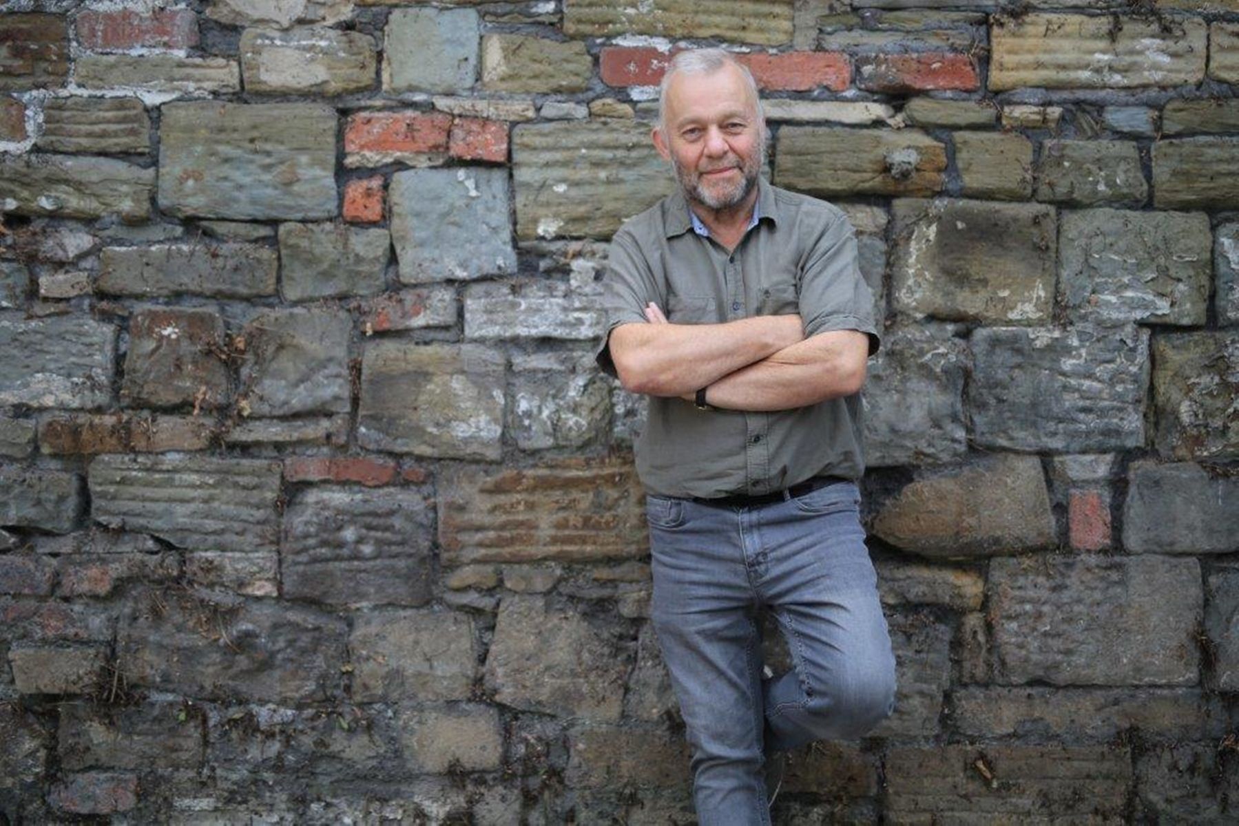 A person wearing a dark patterned shirt stands at a microphone, gesturing with both hands while speaking.