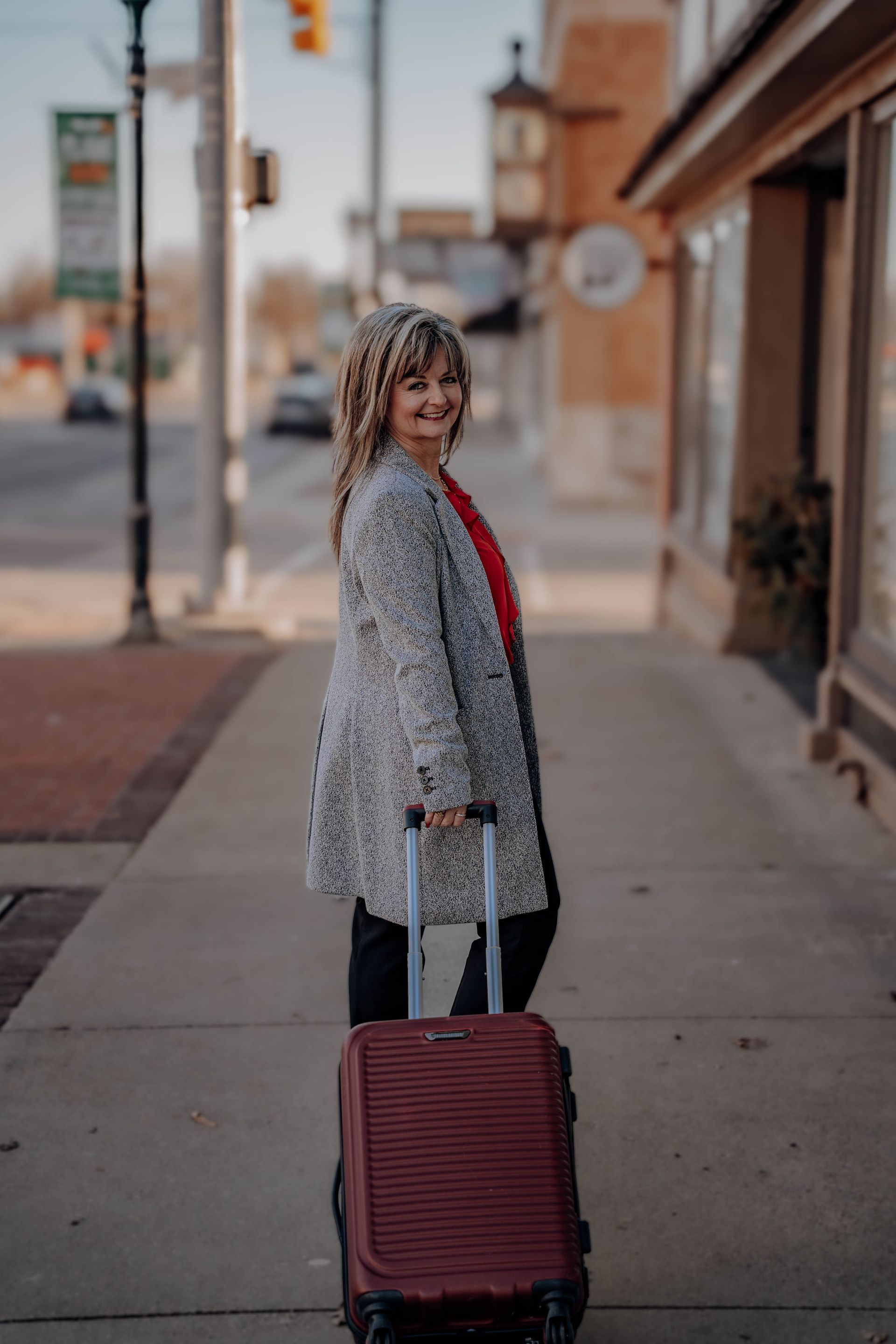 Shelia in a patterned coat and red shirt walks on a sidewalk, pulling a red rolling suitcase.