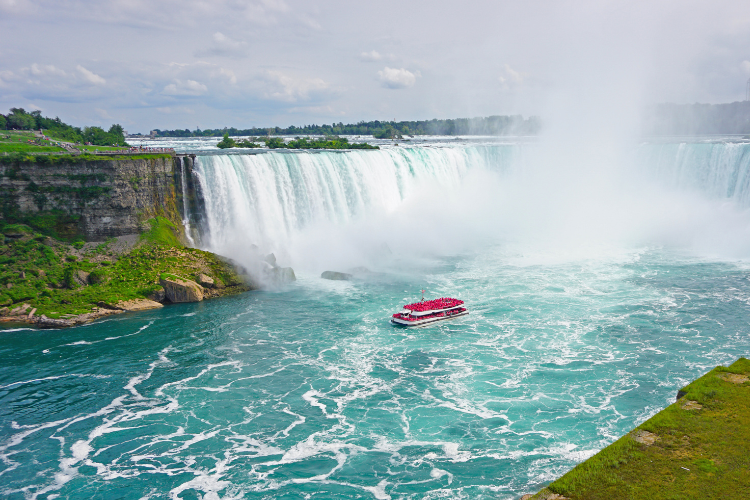 A red tourist boat navigates the turbulent turquoise water at the base of the massive, mist-covered Horseshoe Falls.