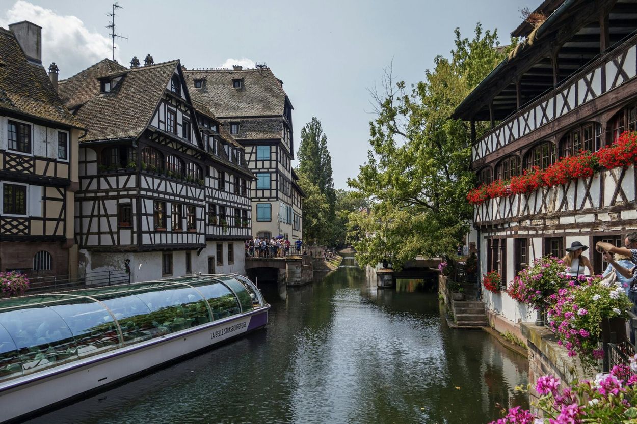 A narrow canal flows between historic half-timbered buildings in Strasbourg, France, with a tour boat in the water.
