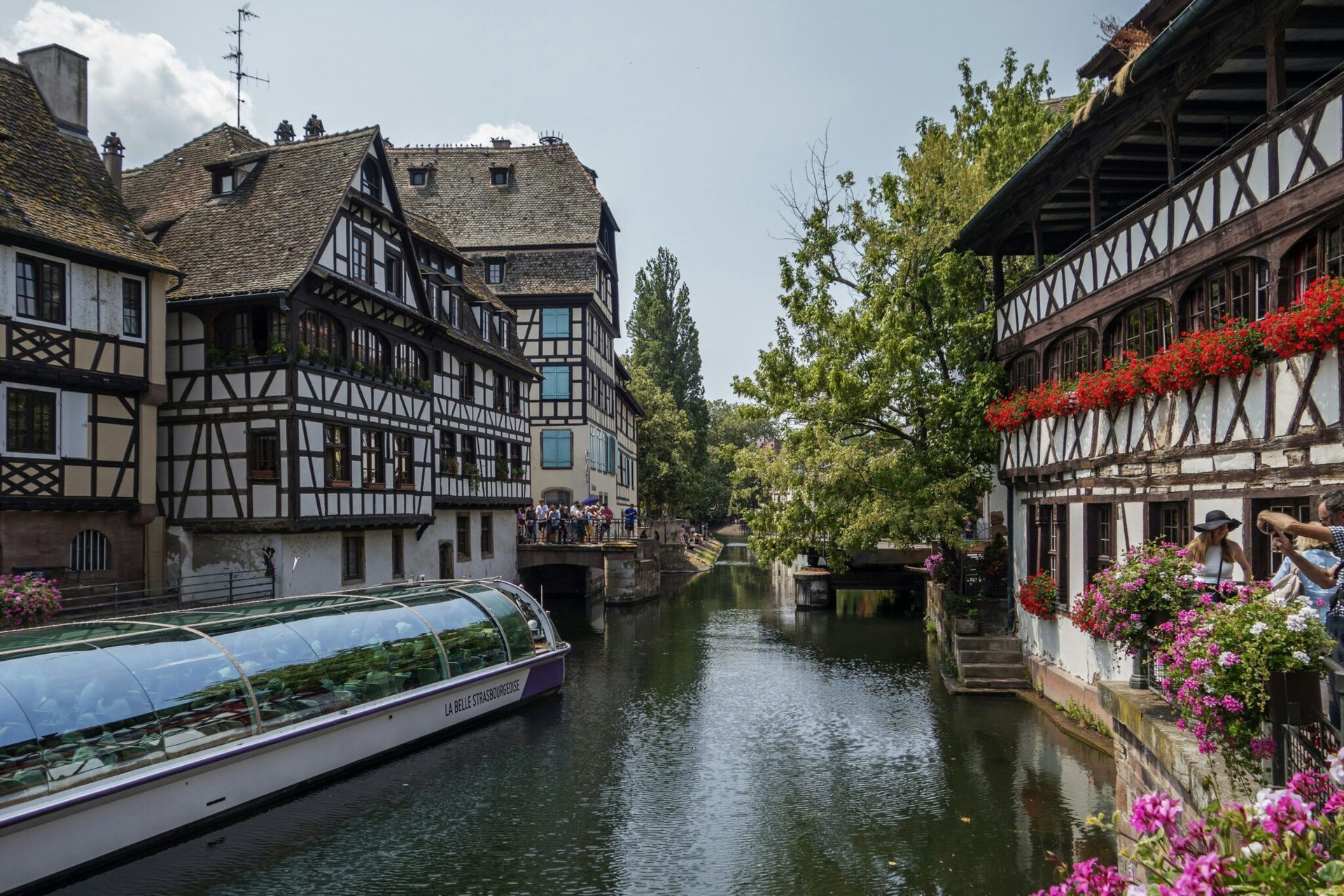 A narrow canal flows between historic half-timbered buildings in Strasbourg, France, with a tour boat in the water.
