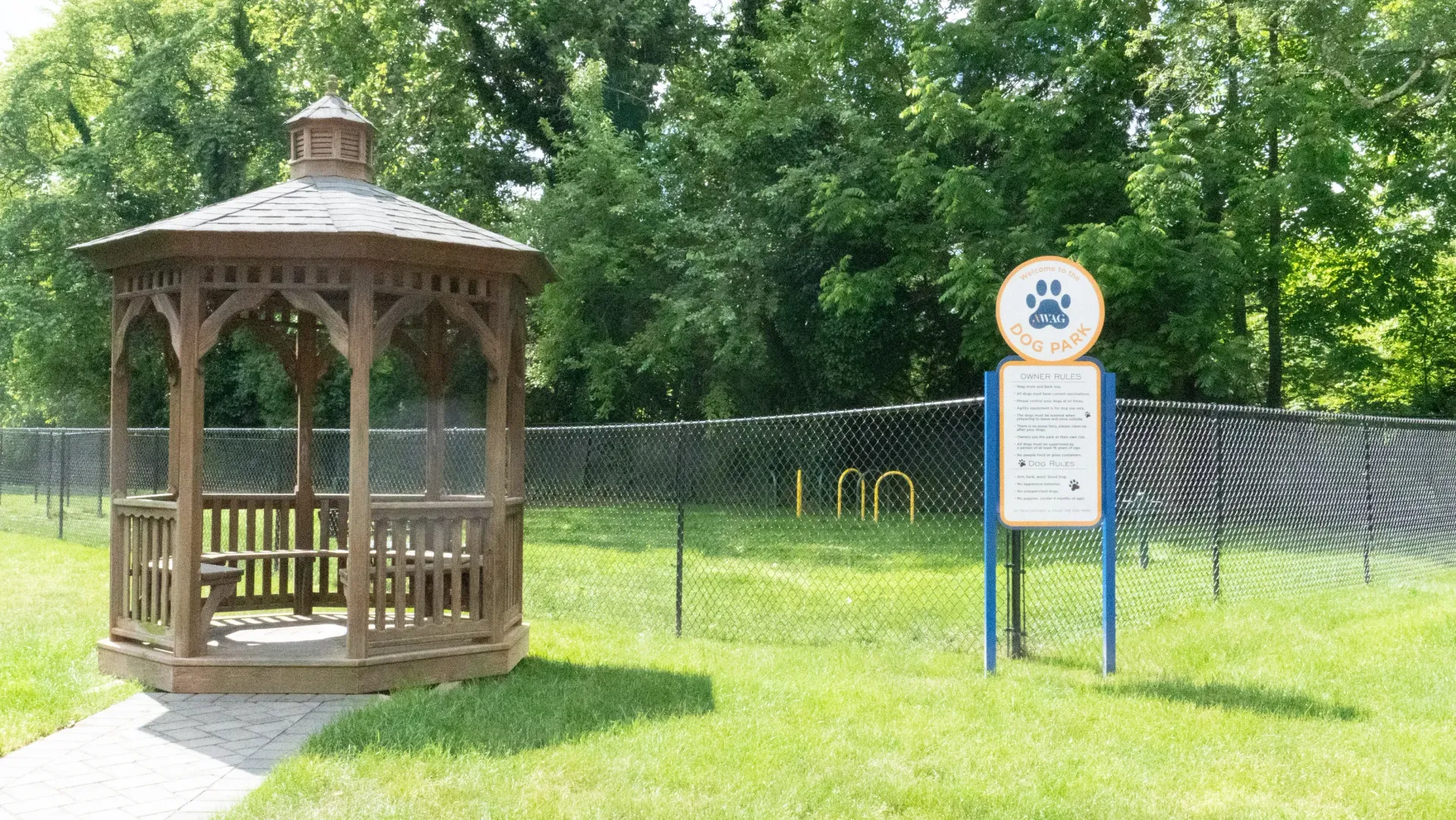 Wooden gazebo by a chain-link fence with a dog park sign in a grassy area.
