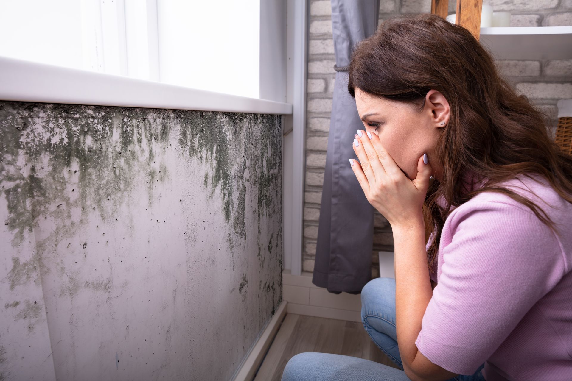 A woman is covering her mouth with her hands while looking at a moldy wall.