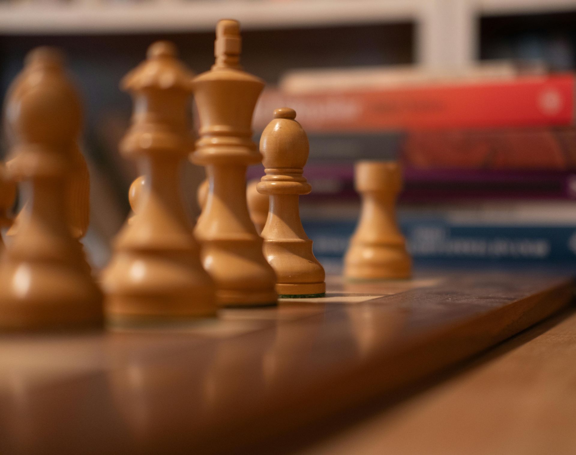 Chess pieces on a wooden board, with blurred books in the background.
