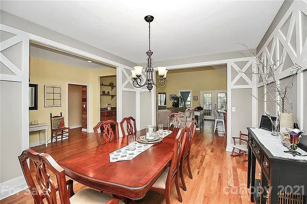 Dining room with large wooden table, chairs, and chandelier. Hardwood floor, open to kitchen.