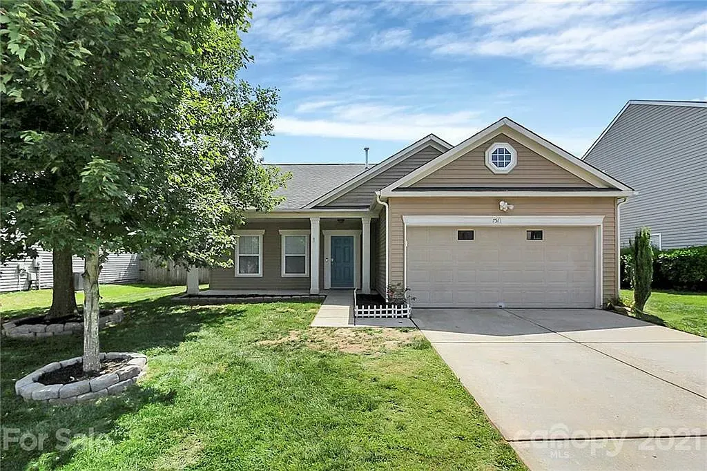 Tan house with a blue door, attached garage, and overgrown lawn under a blue sky.
