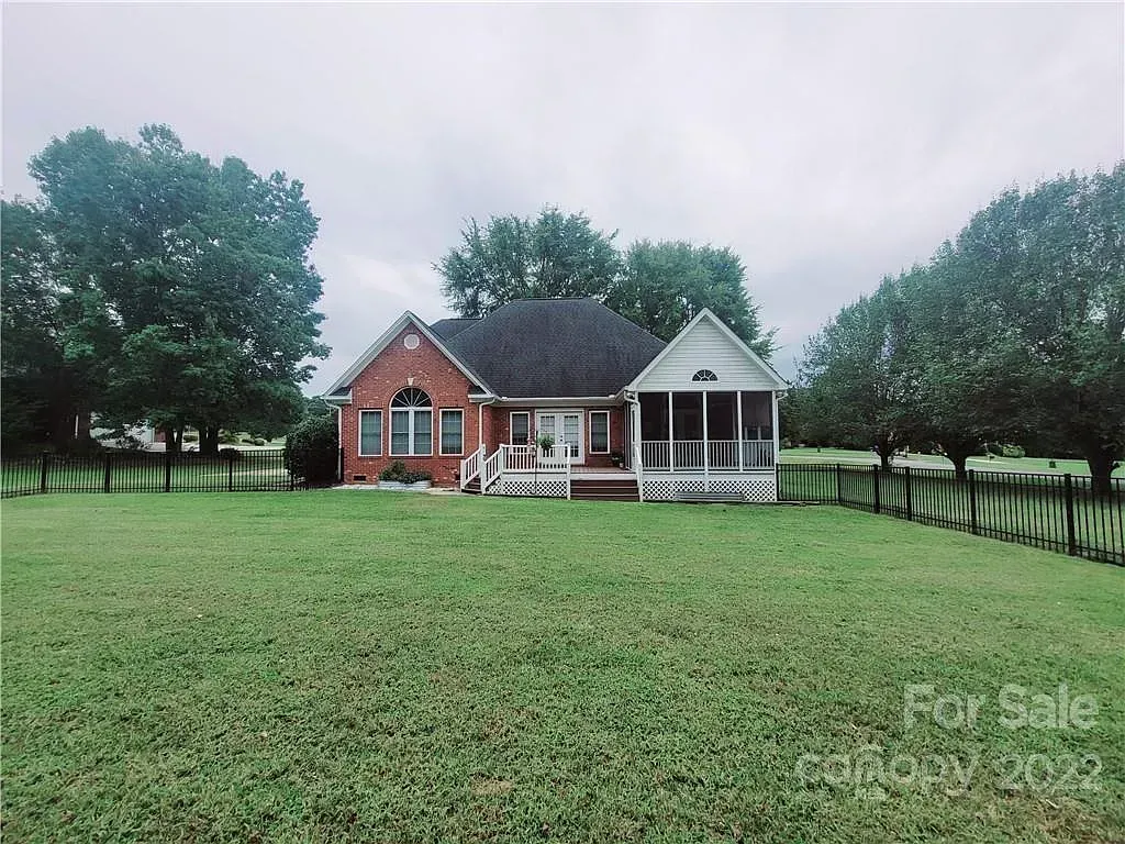 A brick house with a screened porch sits on a grassy lot under a cloudy sky, trees frame the sides.