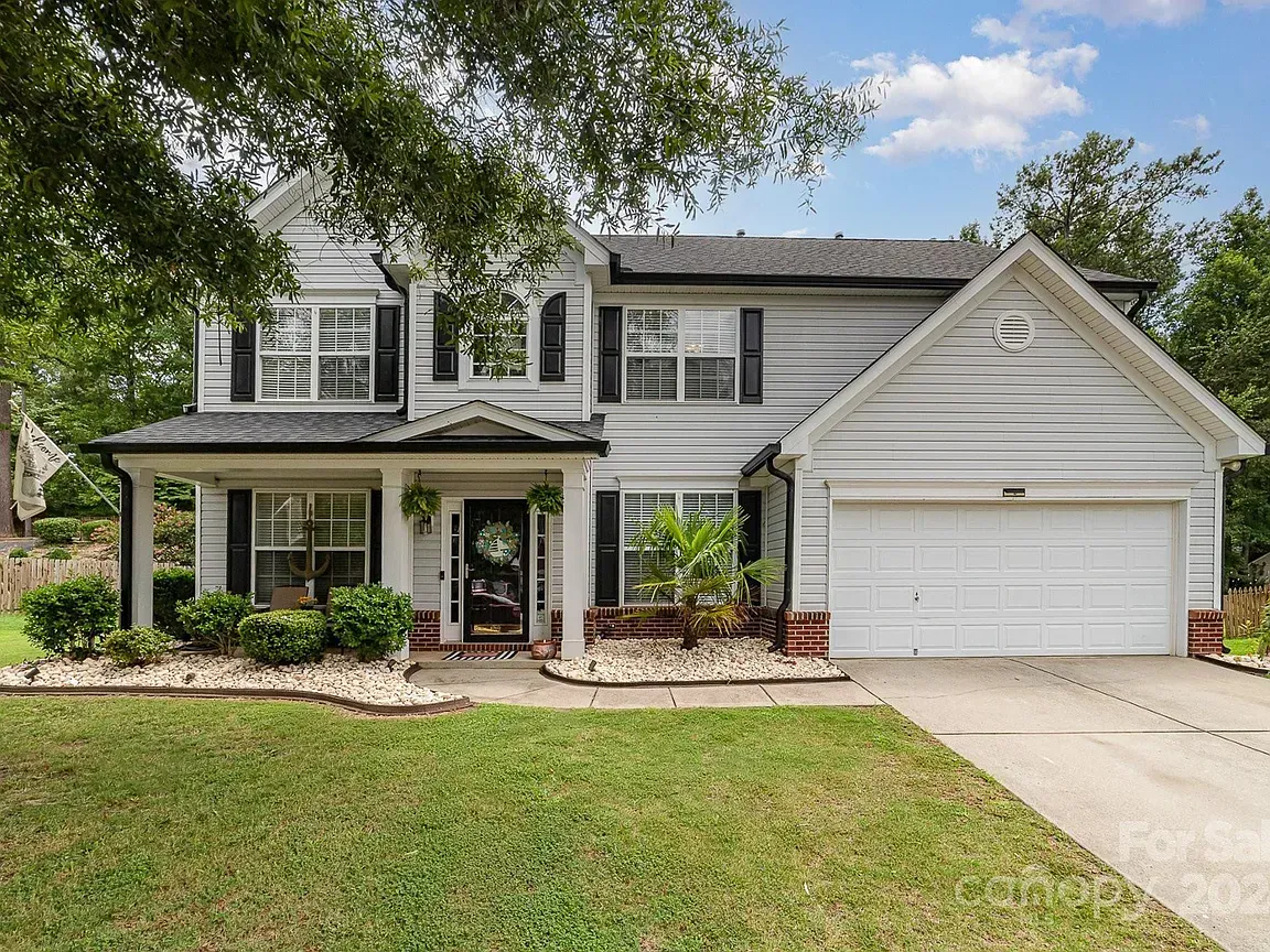 Two-story house with white siding, black shutters, a porch, and a two-car garage. Green lawn and cloudy sky.