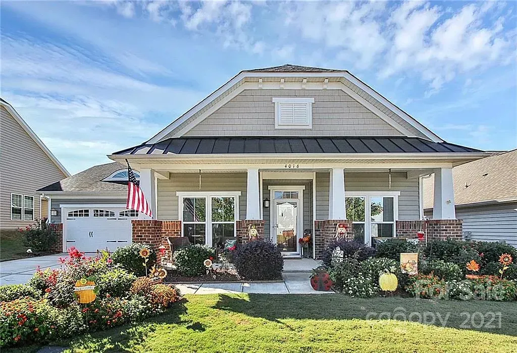 Tan Craftsman-style house with white trim, a brick porch, and landscaping under a blue sky.