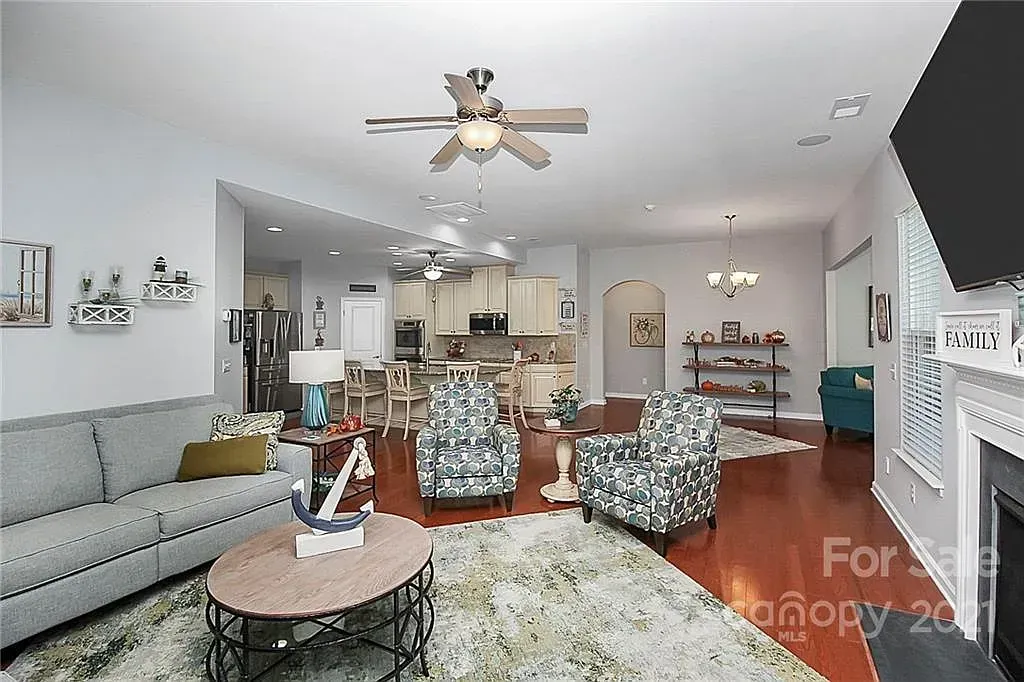 Living room with open layout to kitchen and dining. Wood floors, neutral walls, couches, chairs, fireplace.