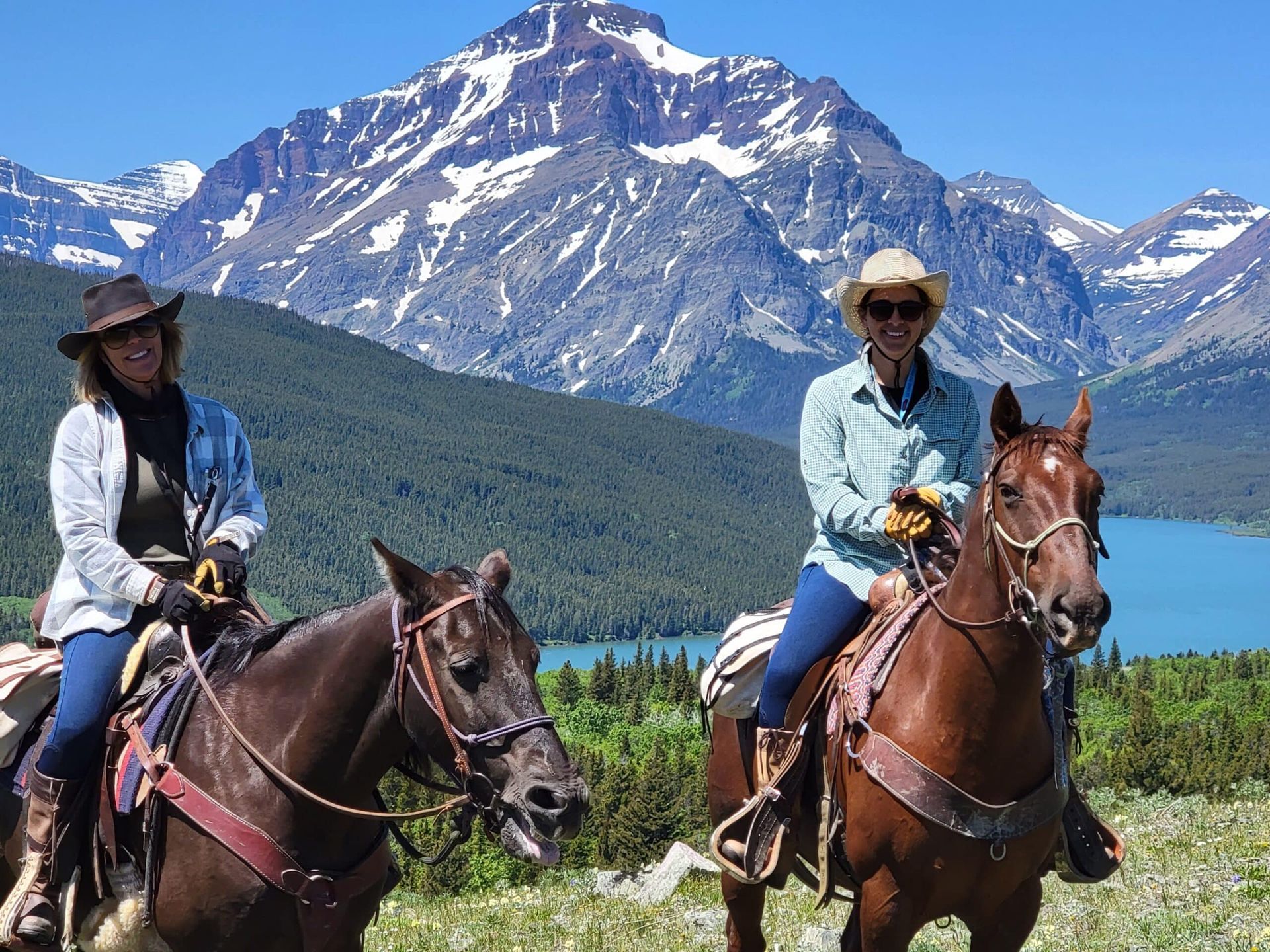 Two women are riding horses in front of mountains and a lake.